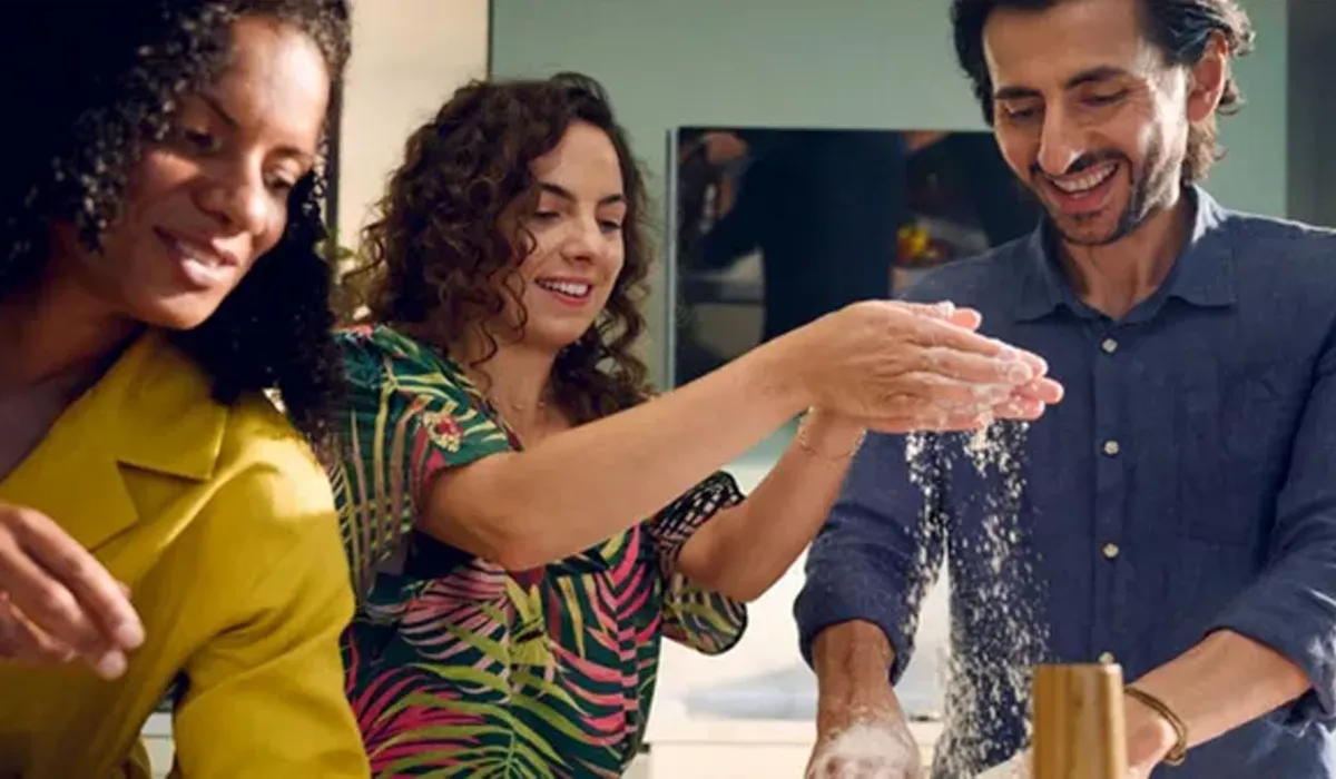 Three smiling people are in a kitchen, engaged in a playful cooking activity. A woman with curly hair, wearing a tropical-patterned shirt, claps her flour-dusted hands together, sending a cloud of flour onto the hands and blue shirt of a man next to her, who is also smiling broadly. Another woman in a yellow top stands to the left, observing with a smile. The scene conveys a joyful and collaborative atmosphere.