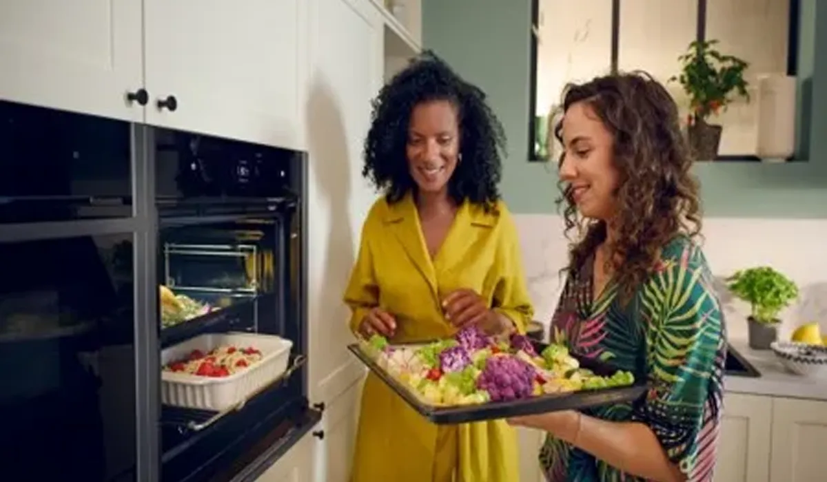 Two smiling women are in a modern kitchen. The woman on the right, with curly hair and wearing a colorful tropical-patterned shirt, holds a large black baking tray filled with colorful vegetables, including purple and green cauliflower, broccoli, and tomatoes.