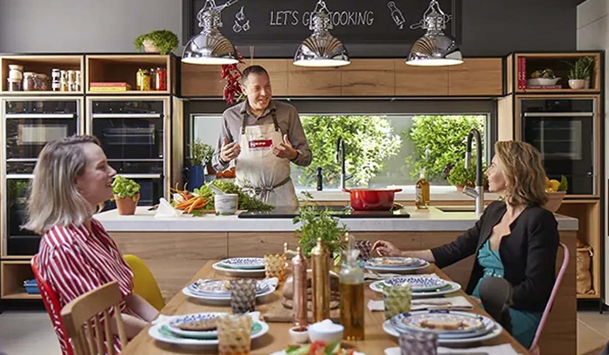 A male chef in an apron stands behind a kitchen island, demonstrating cooking to two women seated at a dining table, with a red pot on an induction hob and fresh ingredients around him. The modern kitchen features built-in ovens, wooden cabinetry, and a chalkboard sign above with "LET'S GO COOKING", creating an engaging and educational atmosphere.