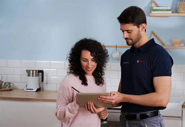 Una persona con un maglione rosa e un'altra con una polo blu scuro discutono e rivedono le informazioni su un tablet in una cucina moderna.