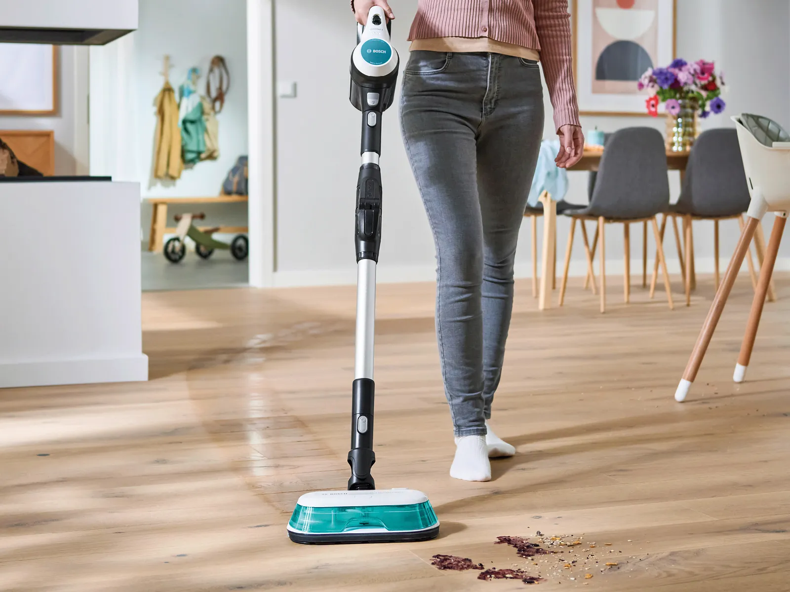 Eine Person in grauer Hose und barfuß reinigt einen Holzboden mit einem Bodenreiniger. Krümel und Schmutz sind auf dem Boden verteilt.