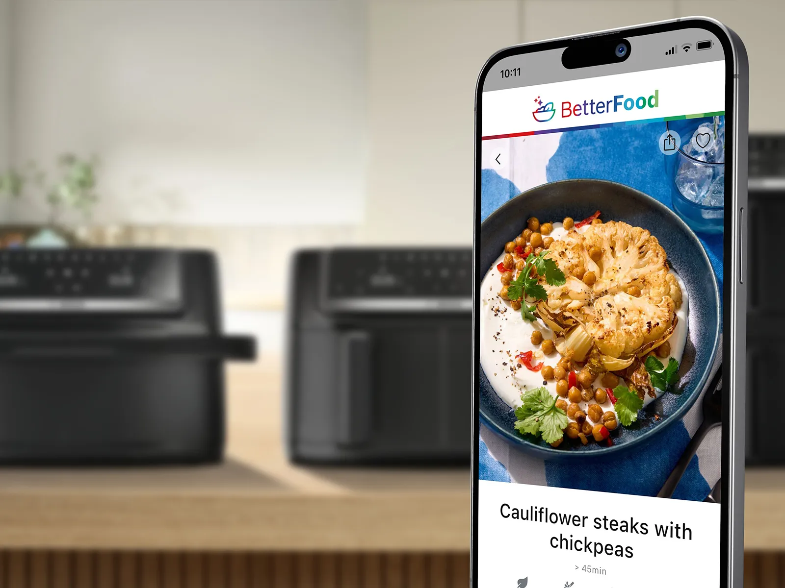 A mobile phone shows the Bosch BetterFood app. In the background there are air fryers on a kitchen counter.