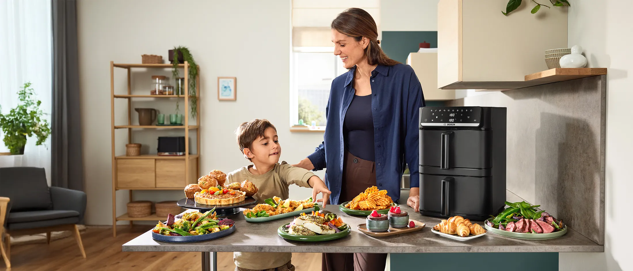 A woman and child stand behind a kitchen counter with the Bosch Series 6 Double Stack Drawer Air Fryer and various air-fried dishes