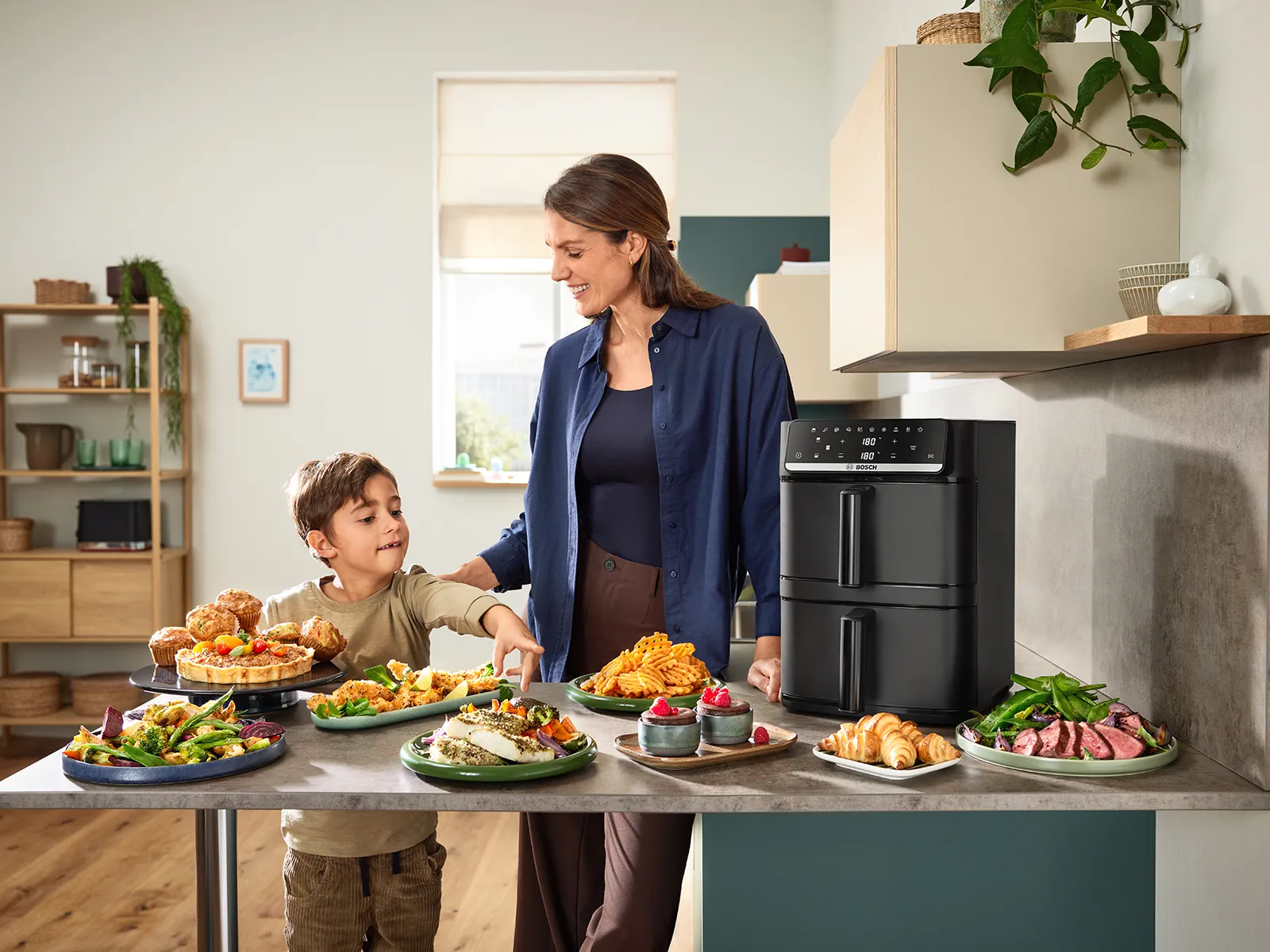 A woman and child stand behind a kitchen counter with the Bosch Series 6 Double Stack Drawer Air Fryer and various air-fried dishes