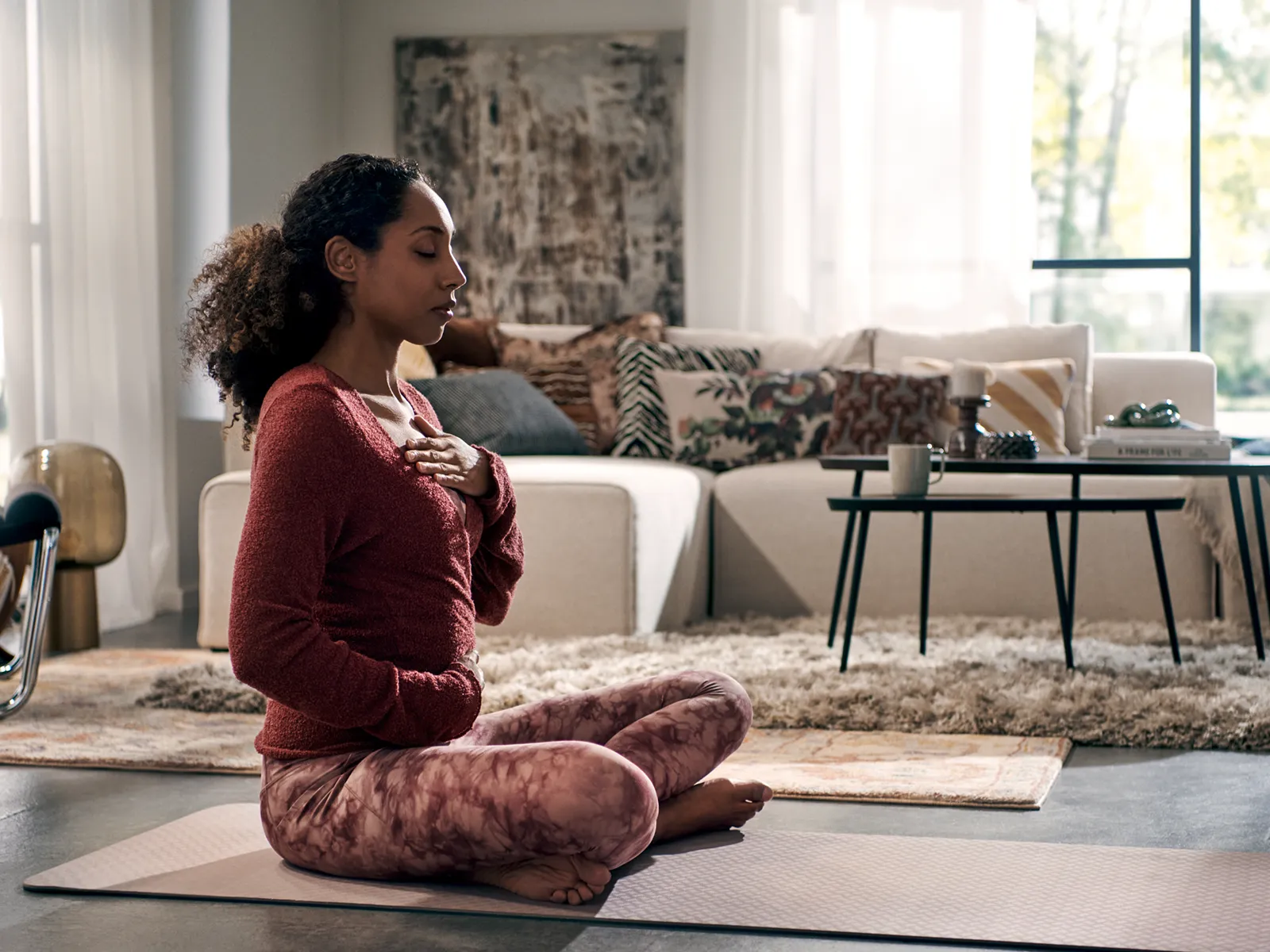 The image shows a woman with curly hair sitting cross-legged on a yoga mat in a cozy living room, appearing to be meditating or practicing mindfulness