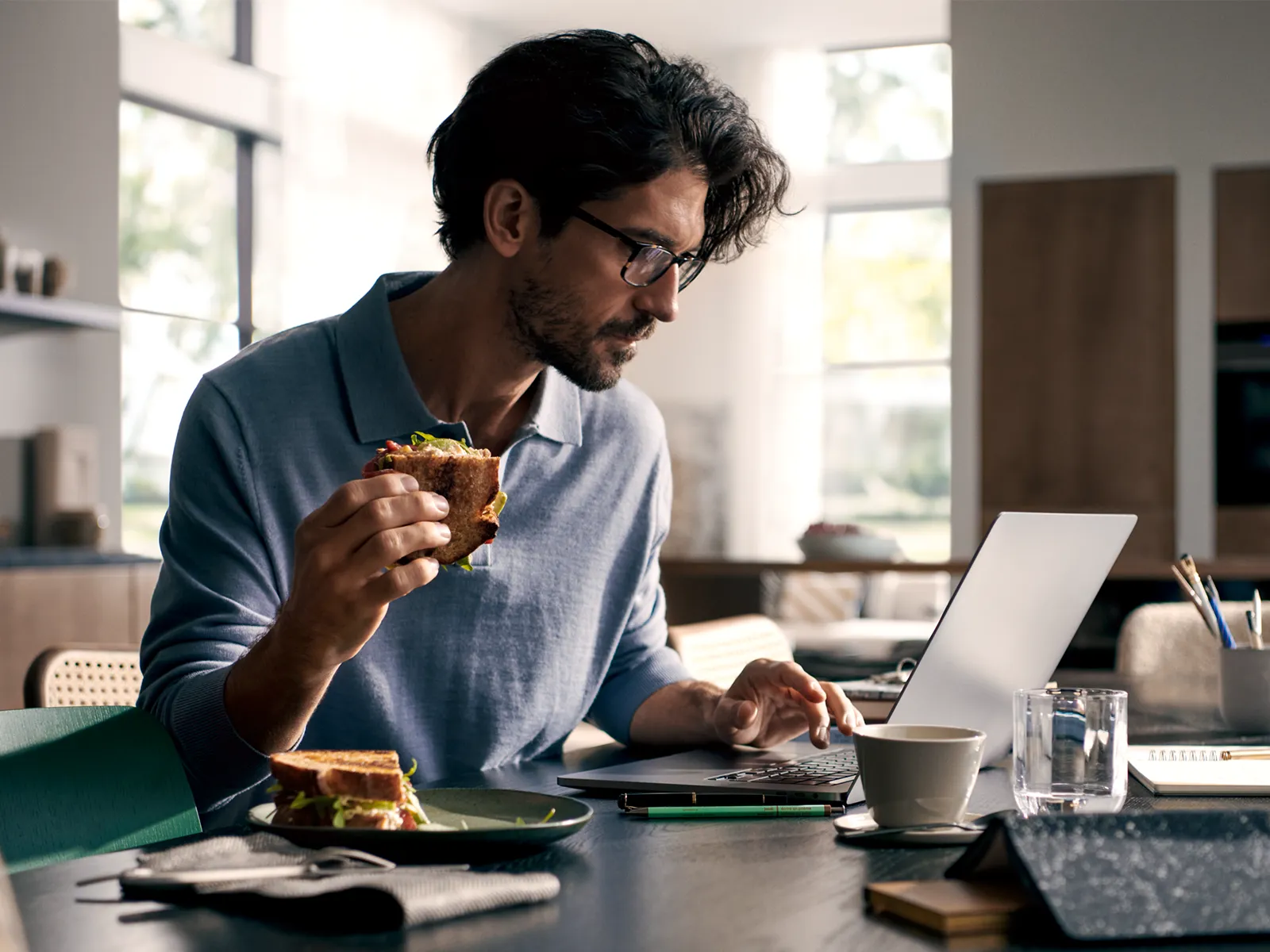 Man in glasses working on laptop, eating snack, surrounded by office supplies on desk.