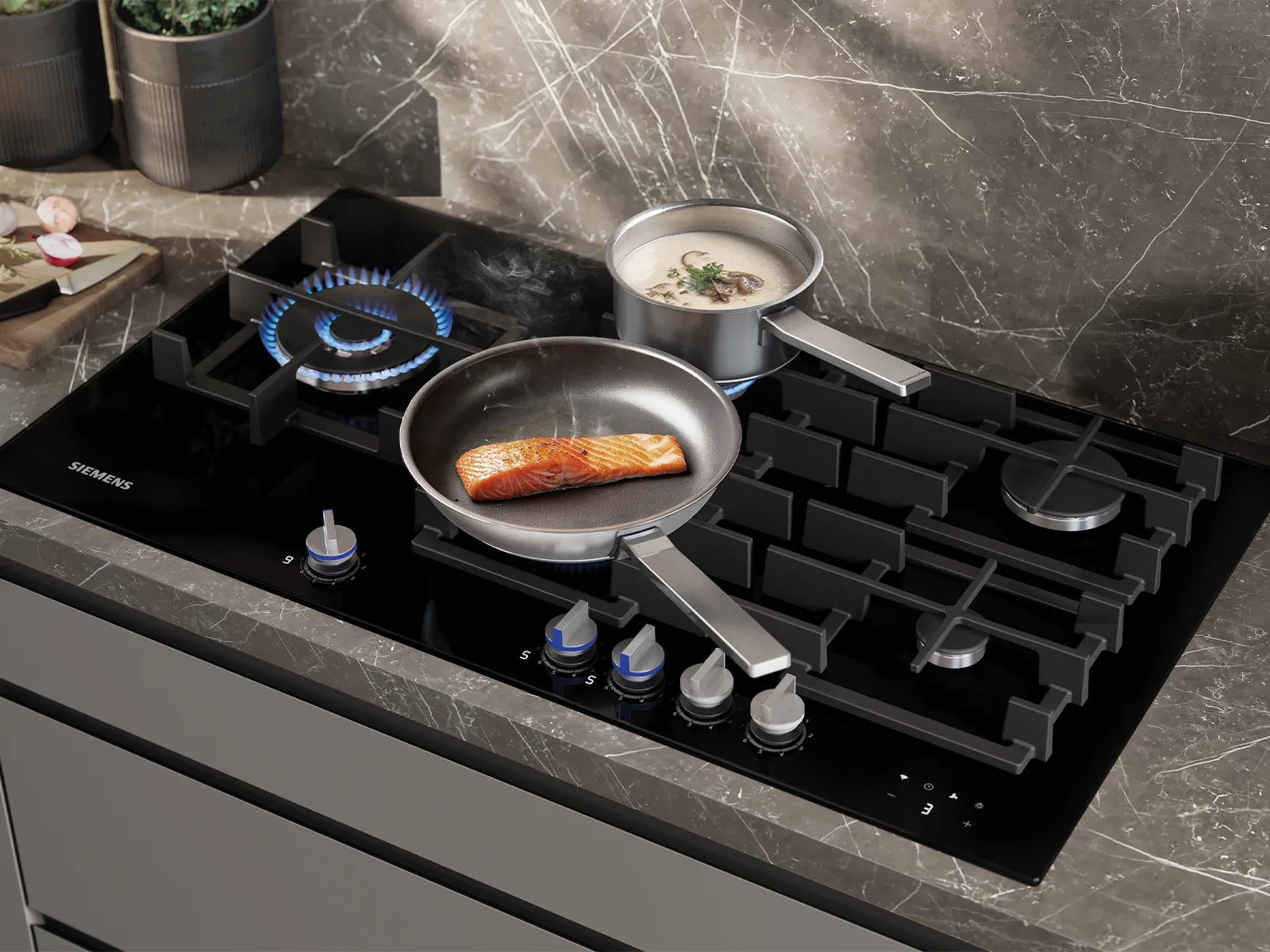A black Siemens gas hob with blue flames, cooking a salmon fillet in a pan and a pot of creamy soup, set on a dark marble countertop.