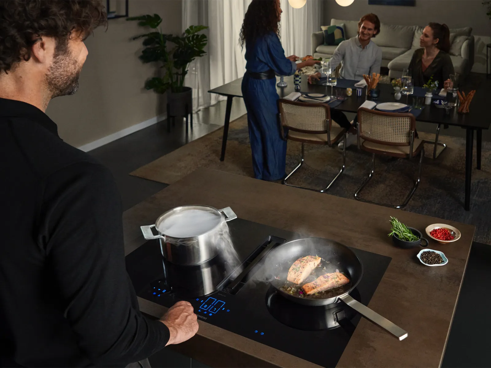 A man cooking salmon and boiling water on an induction hob with an integrated extractor, while friends gather at a dining table in the background.