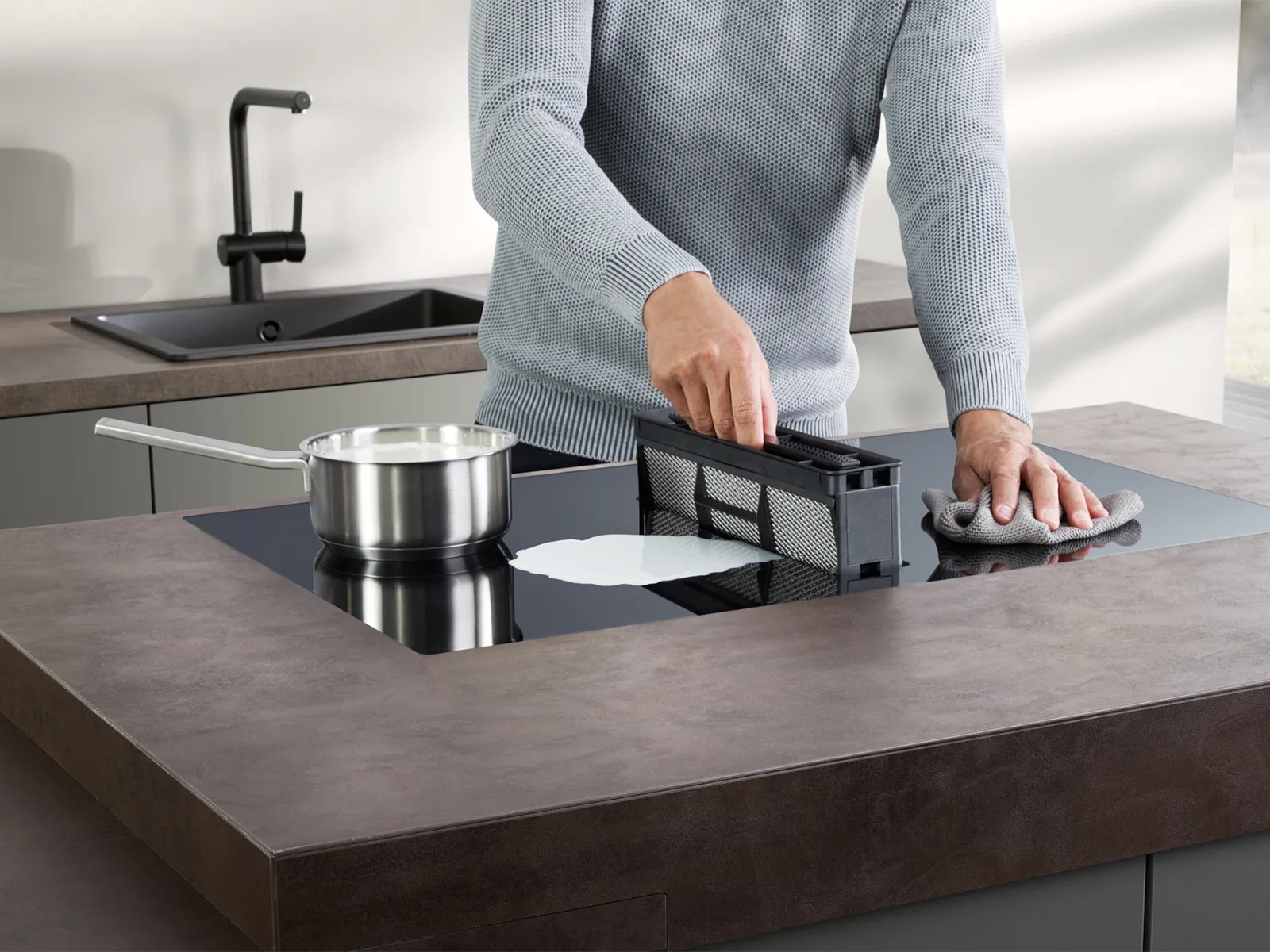 A person cleaning a spill on an induction hob with an integrated downdraft extractor, removing the dishwasher-safe grease filter.