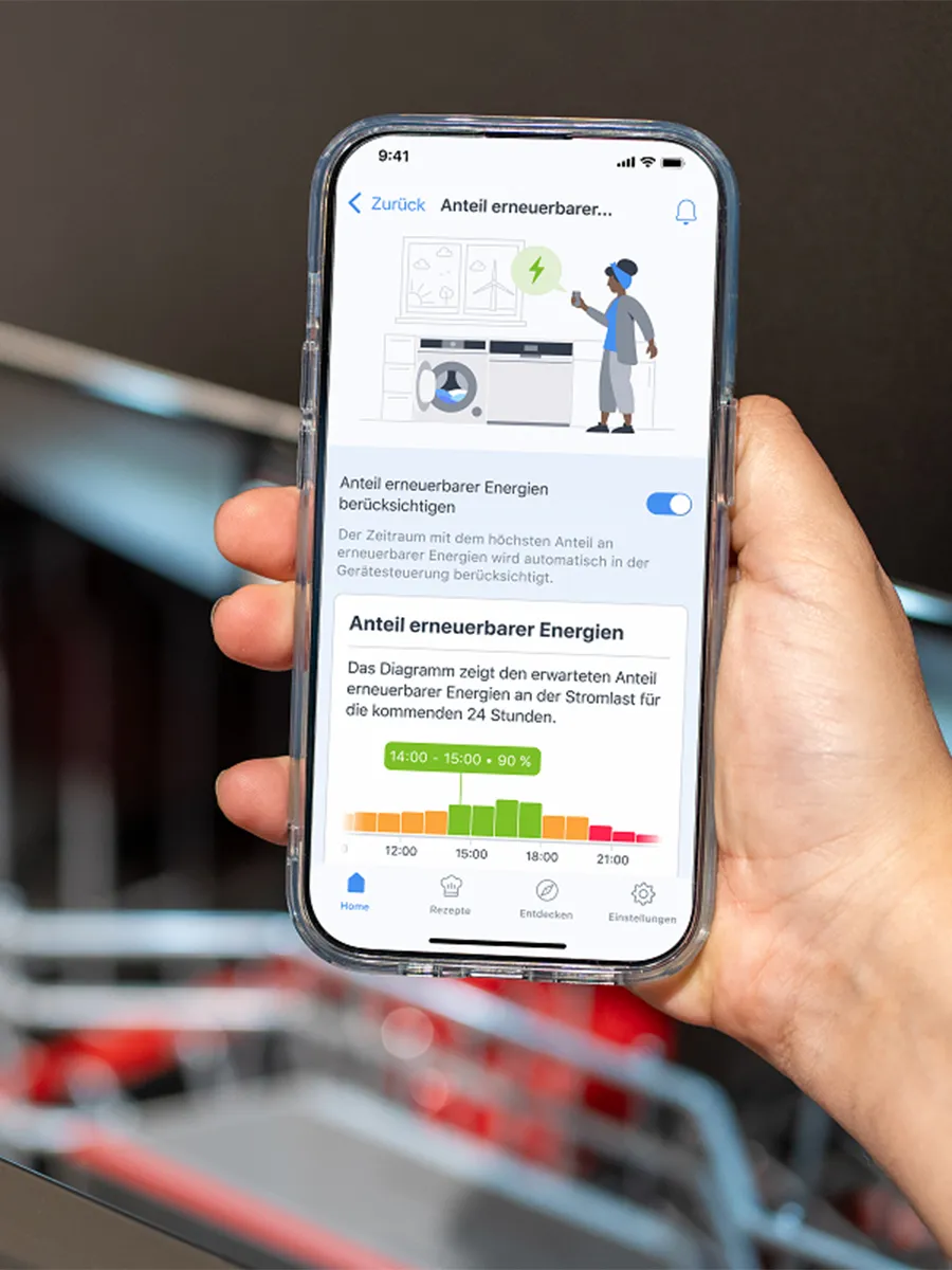 Eine Hand hält ein Smartphone mit der Home Connect App, die den Anteil erneuerbarer Energien für den Betrieb einer Waschmaschine anzeigt.