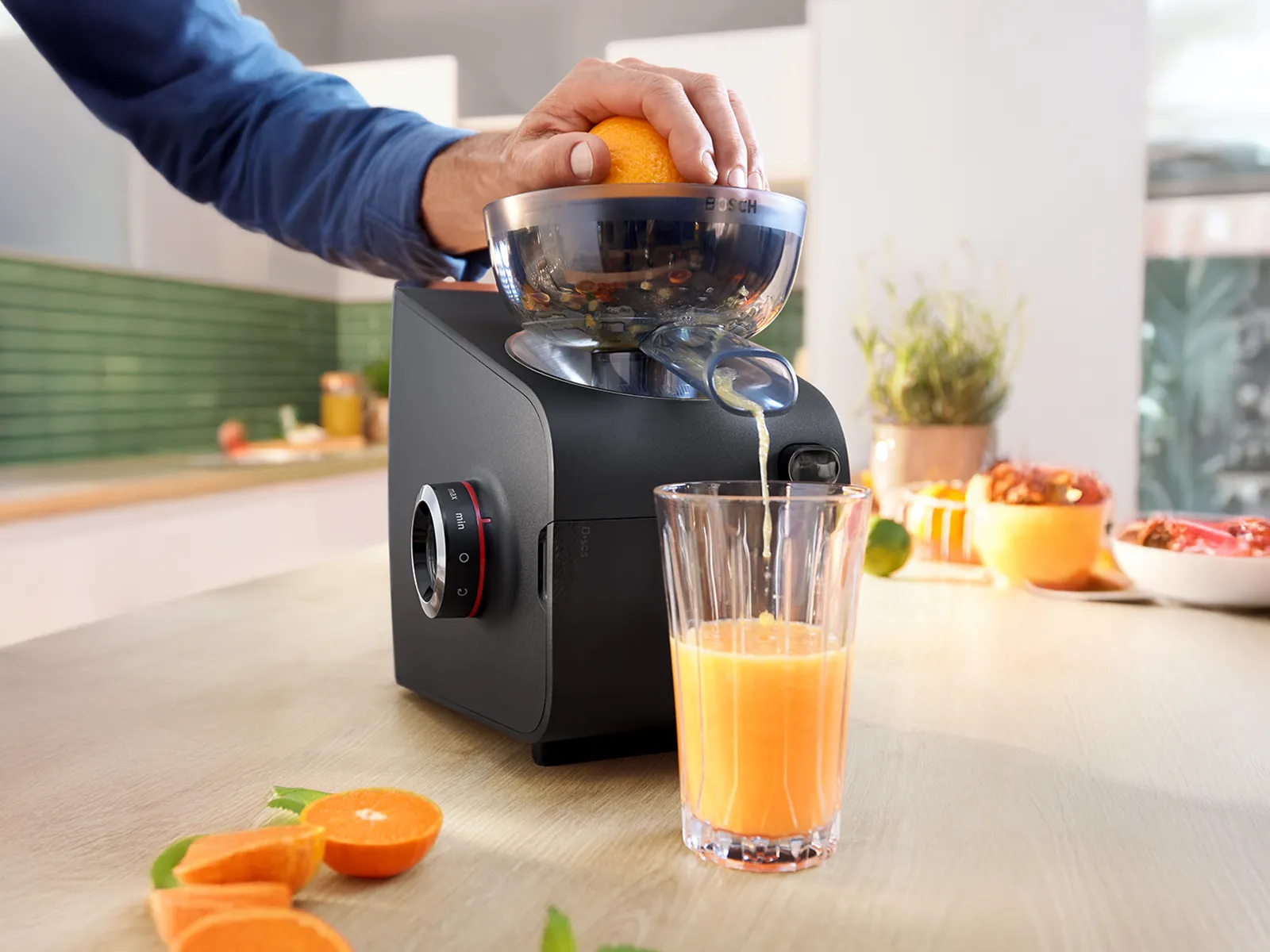 Black electric citrus juicer presses an orange, pouring fresh juice into a tall glass on a kitchen counter.
