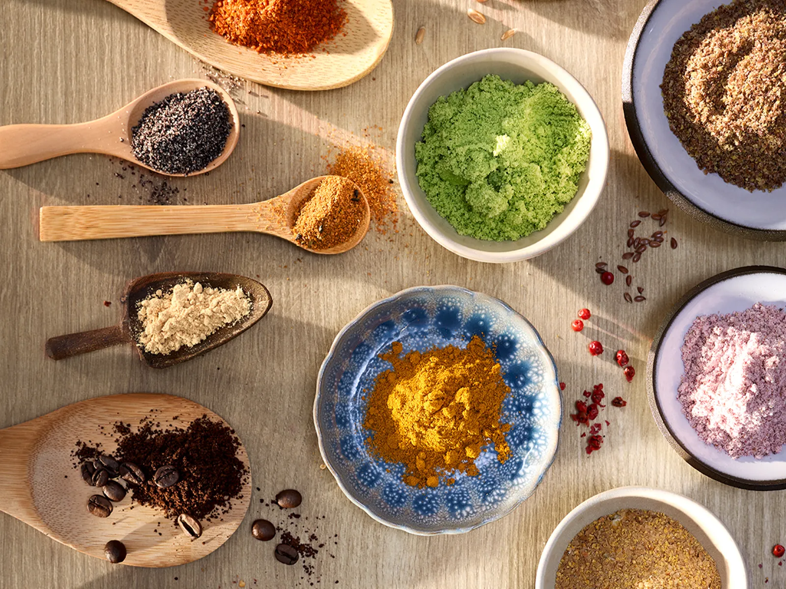 An overhead view of various spices and powders in bowls and spoons, showcasing vibrant colors and diverse textures on a wooden table.