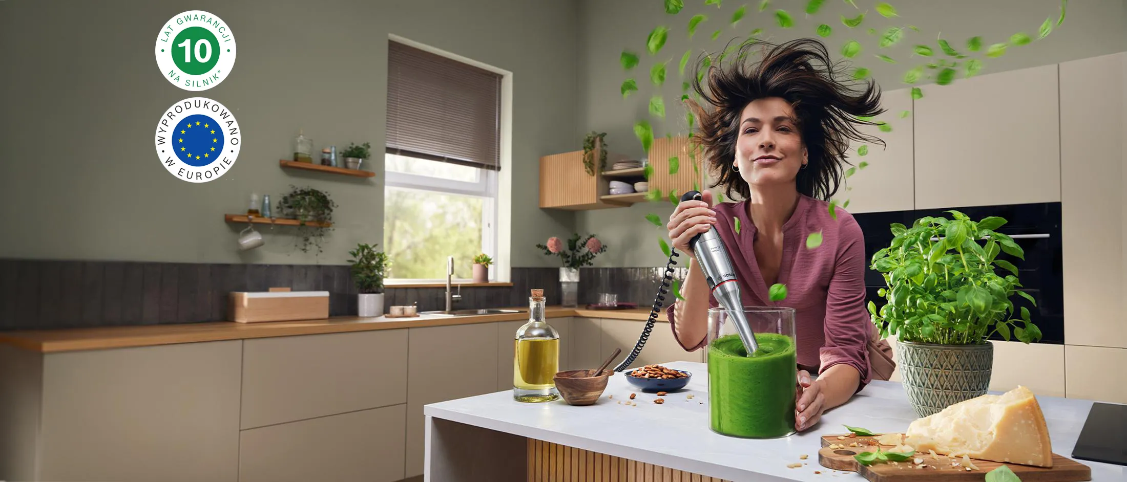 Uśmiechnięta kobieta miksuje zielone smoothie w jasnej kuchni, świeże liście fruwają wokół niej, a w jej otoczeniu są orzechy, ser i bazylia.