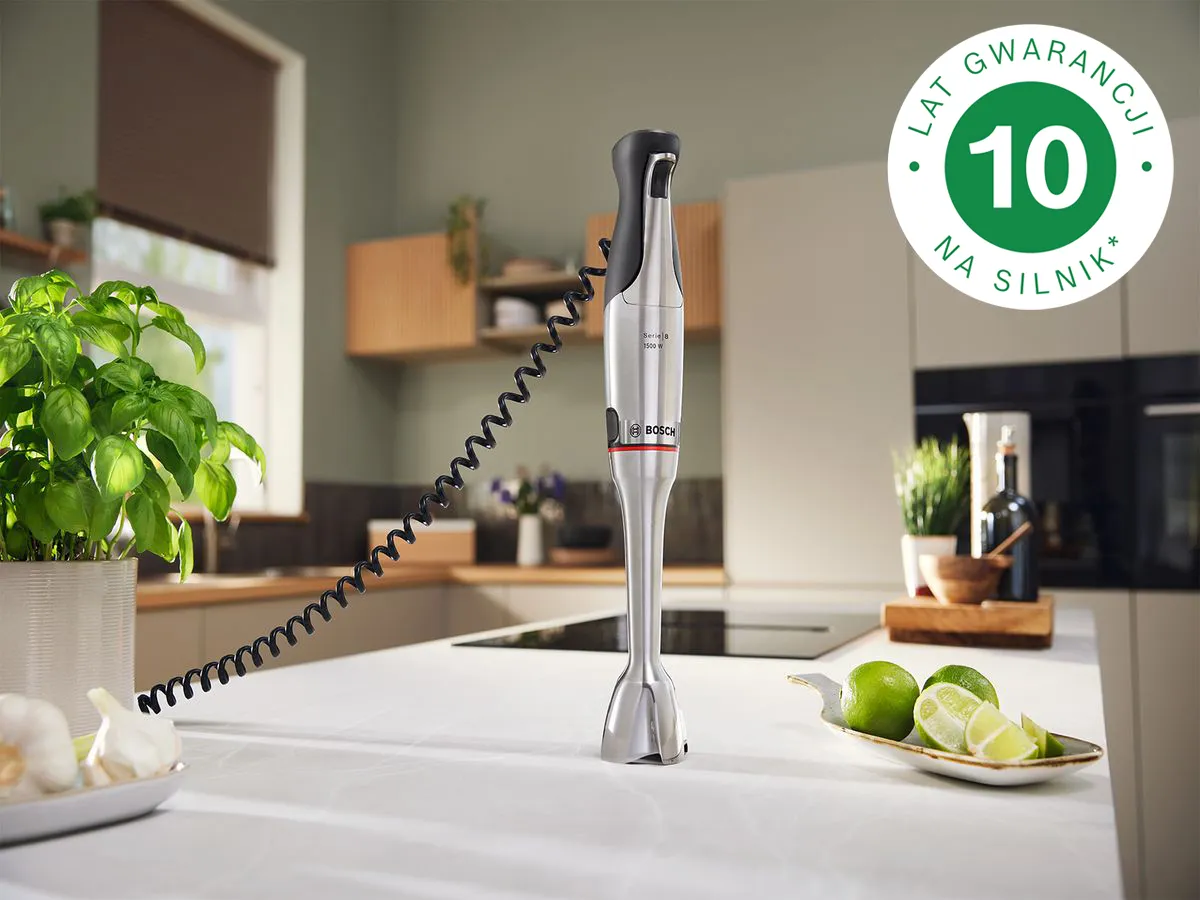 Blender ręczny stoi na blacie kuchennym obok rośliny bazylii, ząbków czosnku i plasterków limonki, w czystej aranżacji kuchennej, z widoczną pieczęcią/logo 10-letniej gwarancji na silnik.