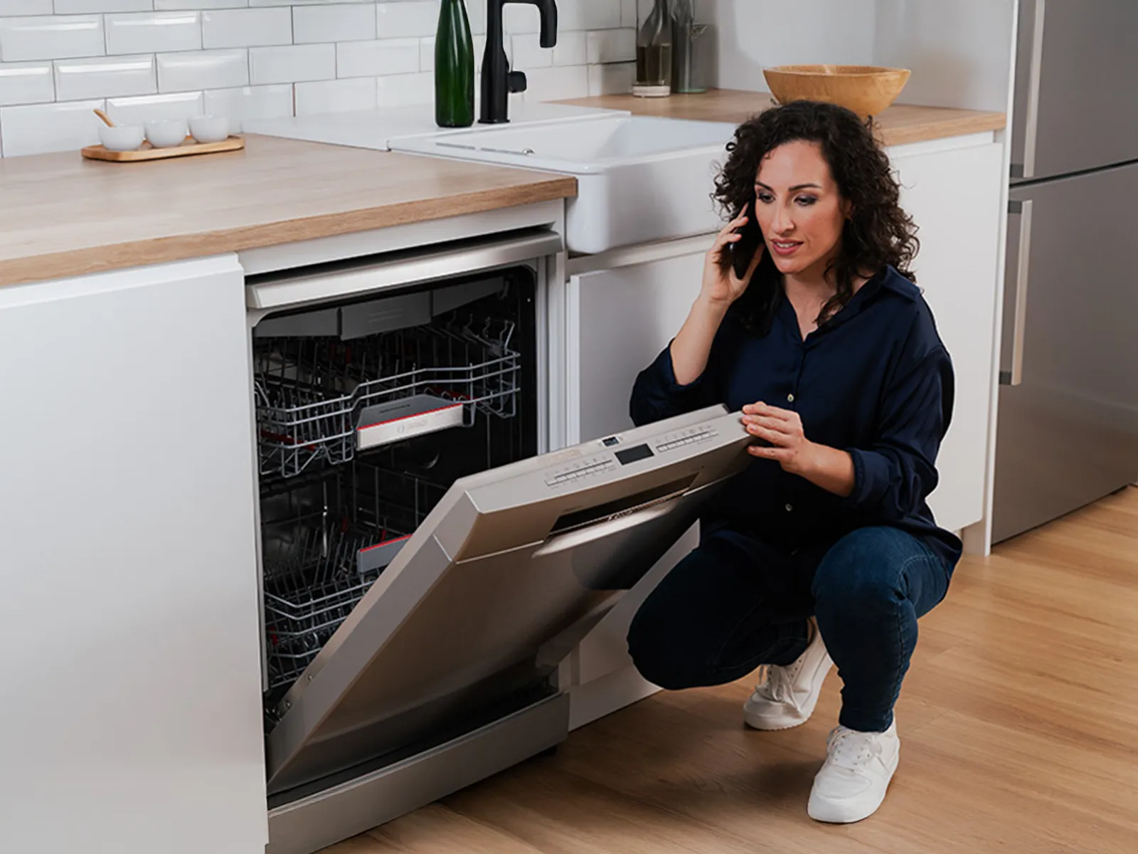 A woman kneeling by an open dishwasher while speaking on the phone in a modern kitchen.