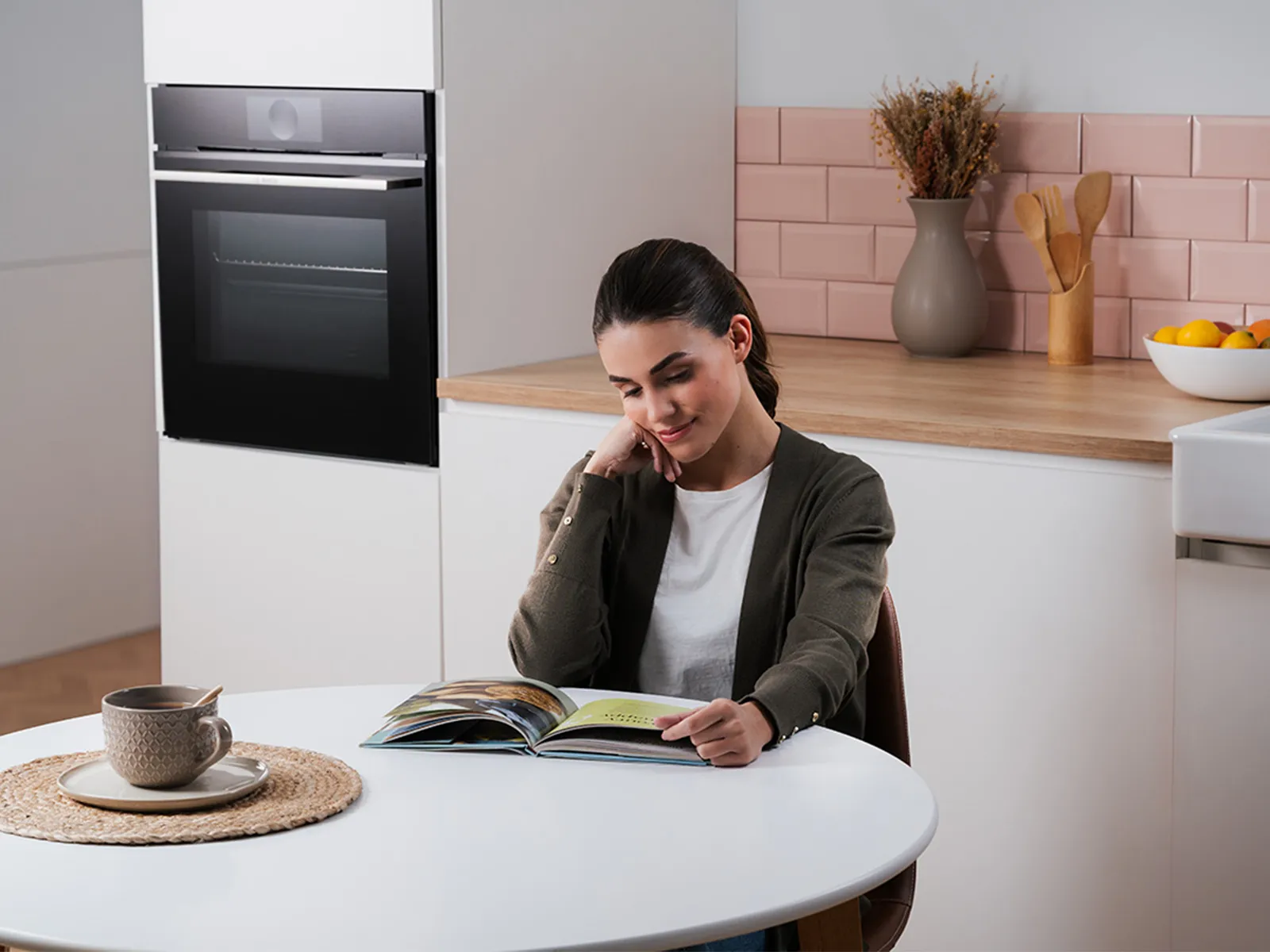 A woman sitting at a kitchen table, reading a book with a cup of tea nearby.