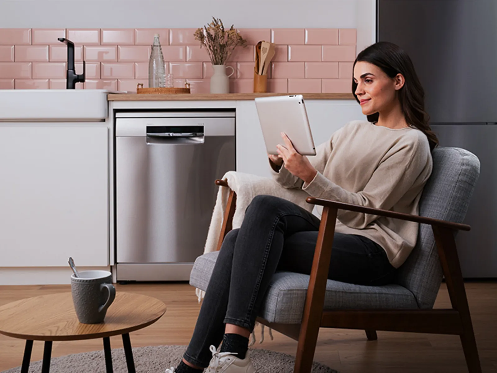 A woman sitting in a modern kitchen-living space, using a tablet while relaxing in an armchair.