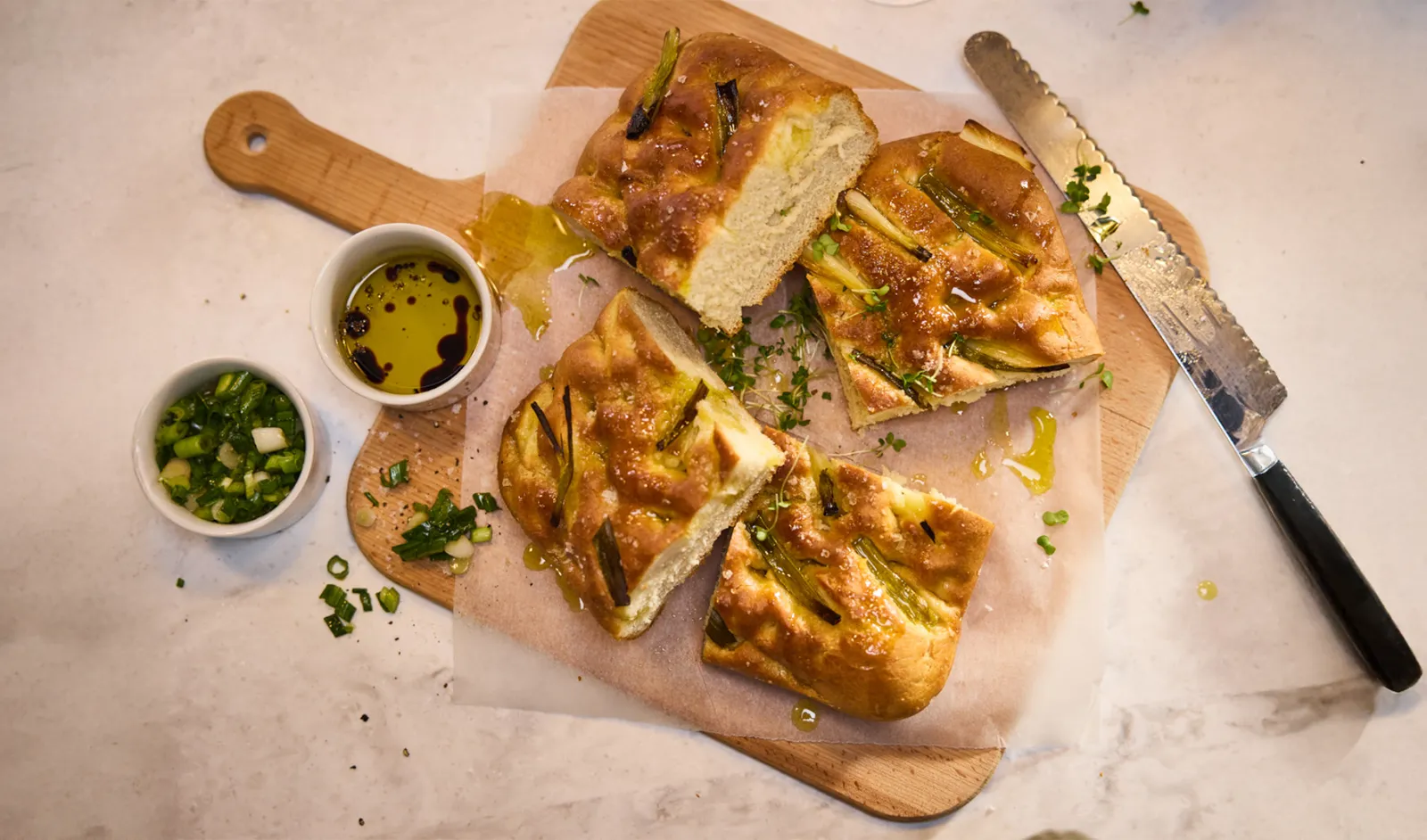 Focaccia fraîchement cuite garnie d'oignons verts, arrosée d'huile d'olive, servie sur une planche à découper en bois avec des sauces.