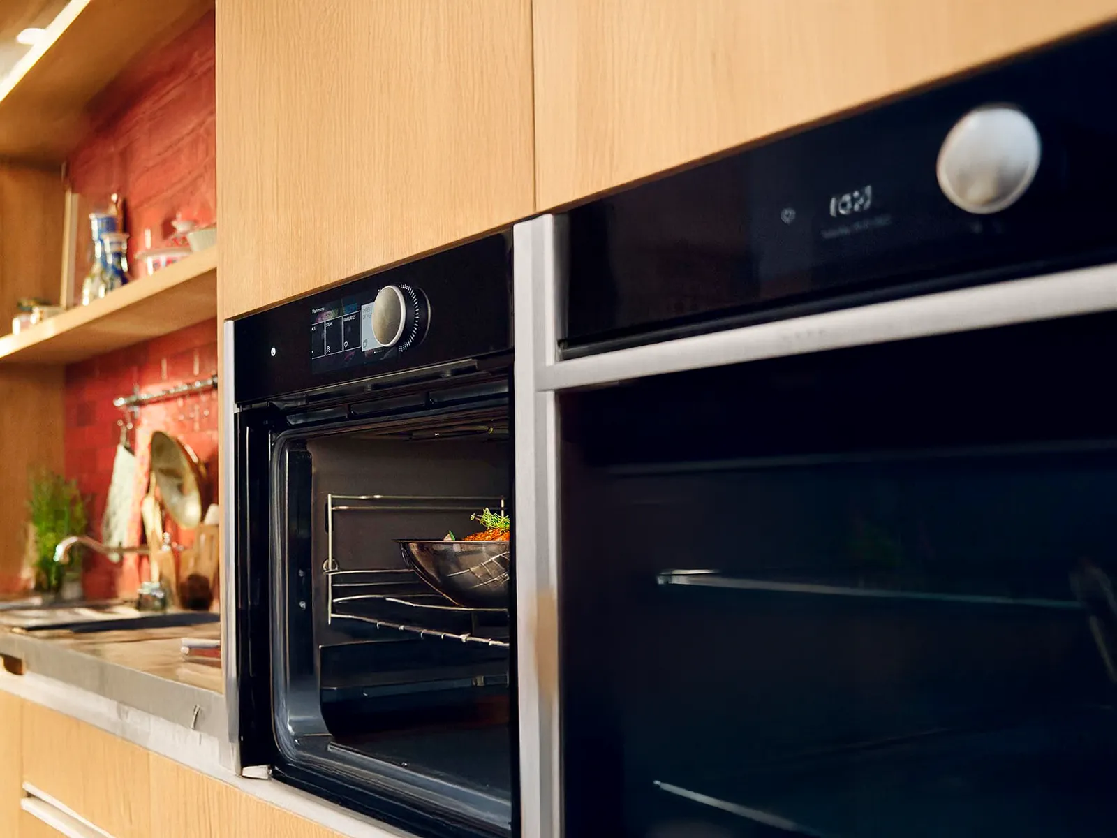 A modern built-in oven with a bowl of food inside, next to another oven, in a kitchen with wooden cabinets.