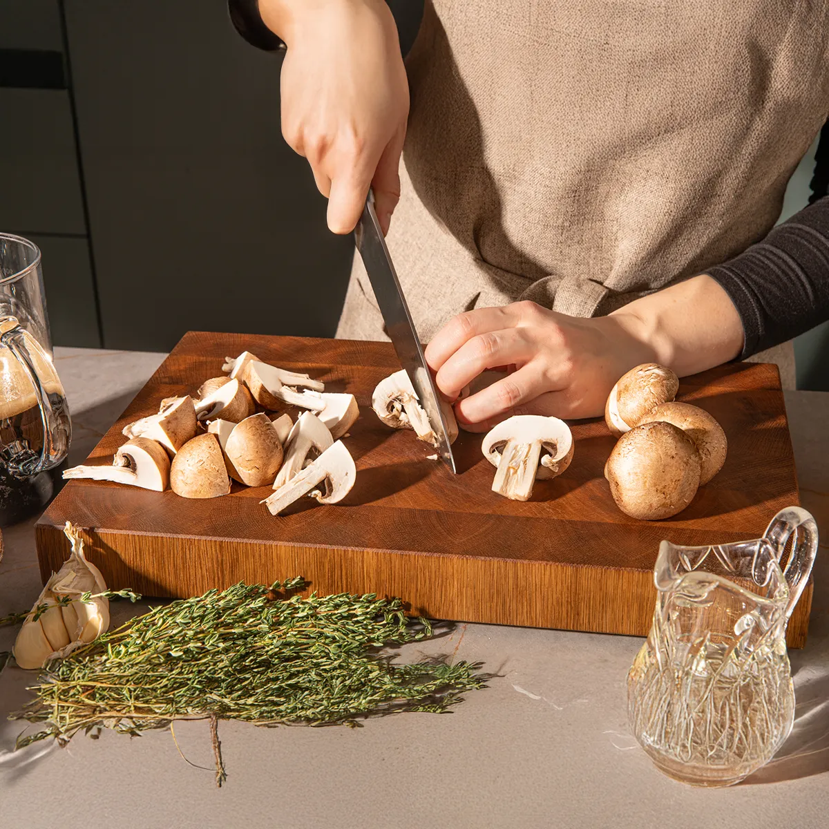 Hands chopping mushrooms on wooden cutting board, surrounded by herbs, wine glasses, and other cooking ingredients.