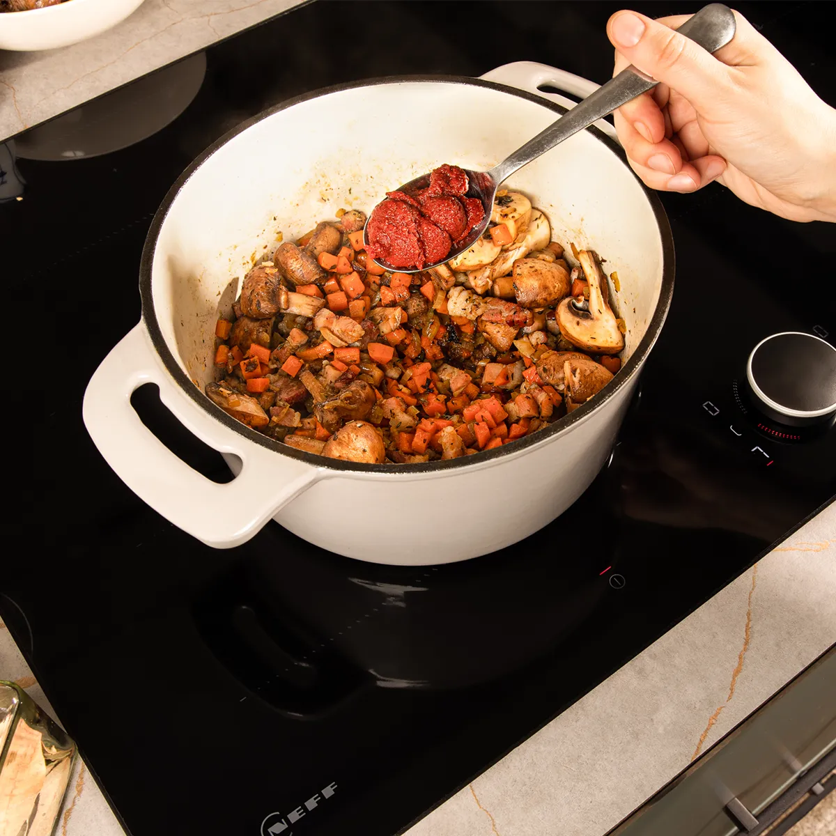 Cooking pot with stew on stove, hand holding spoon, kitchen counter with various cooking ingredients.