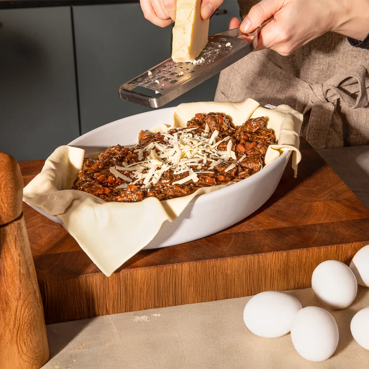Hands grating cheese over a bowl of chili or meat sauce on a wooden cutting board with cooking utensils.