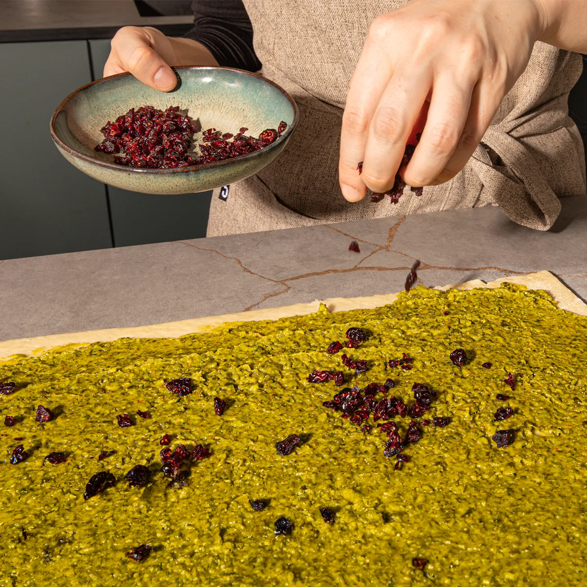 A person sprinkles dried cranberries from a bowl onto a vibrant green dough spread on a countertop.