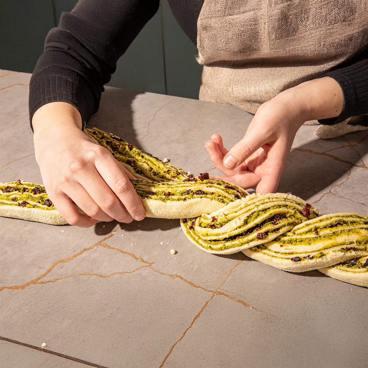 Hands braiding a vibrant green pesto and red cranberry bread on a grey counter, revealing the delicious filling.