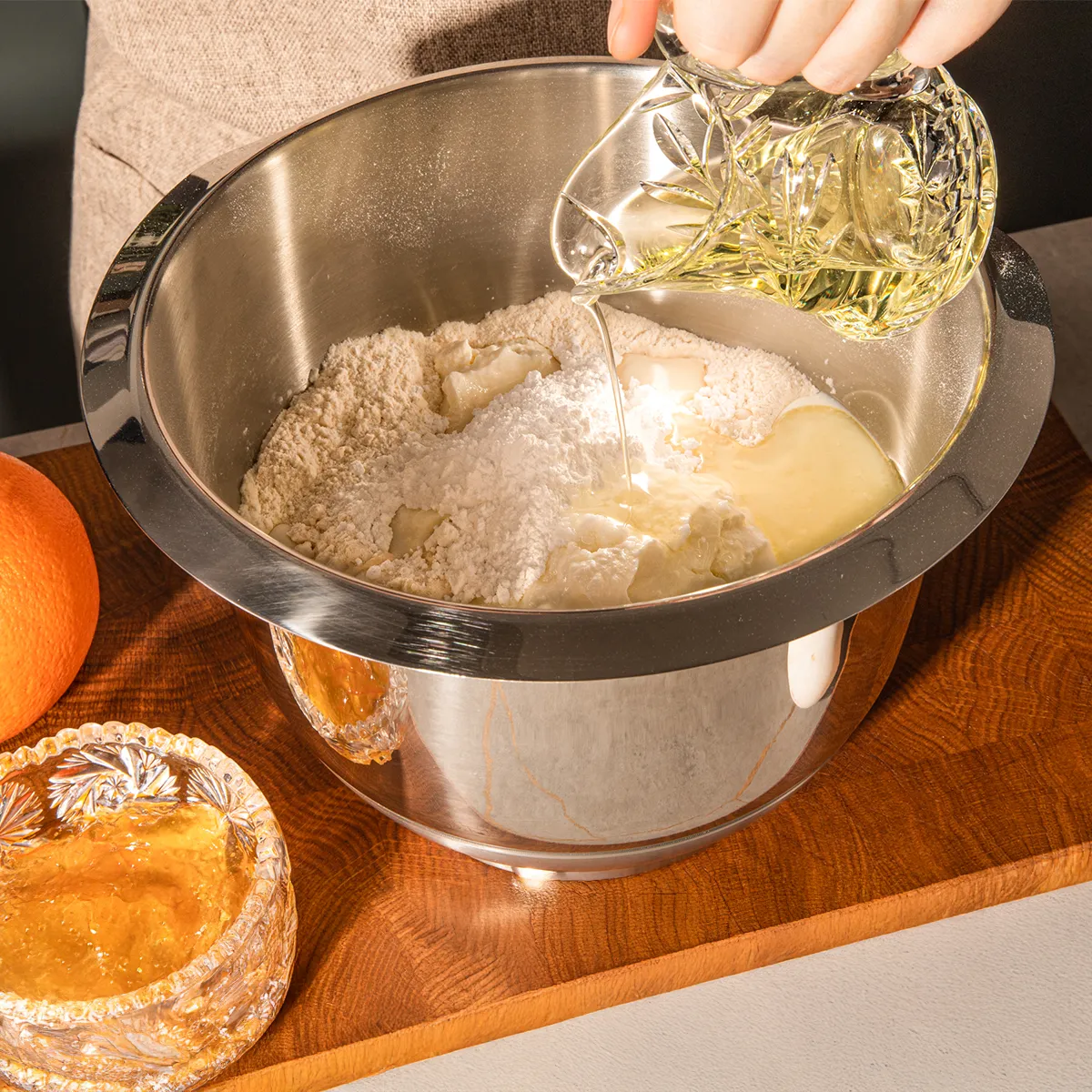 A hand pours oil into a mixing bowl containing flour and other ingredients, with an orange and a small dish of honey nearby.