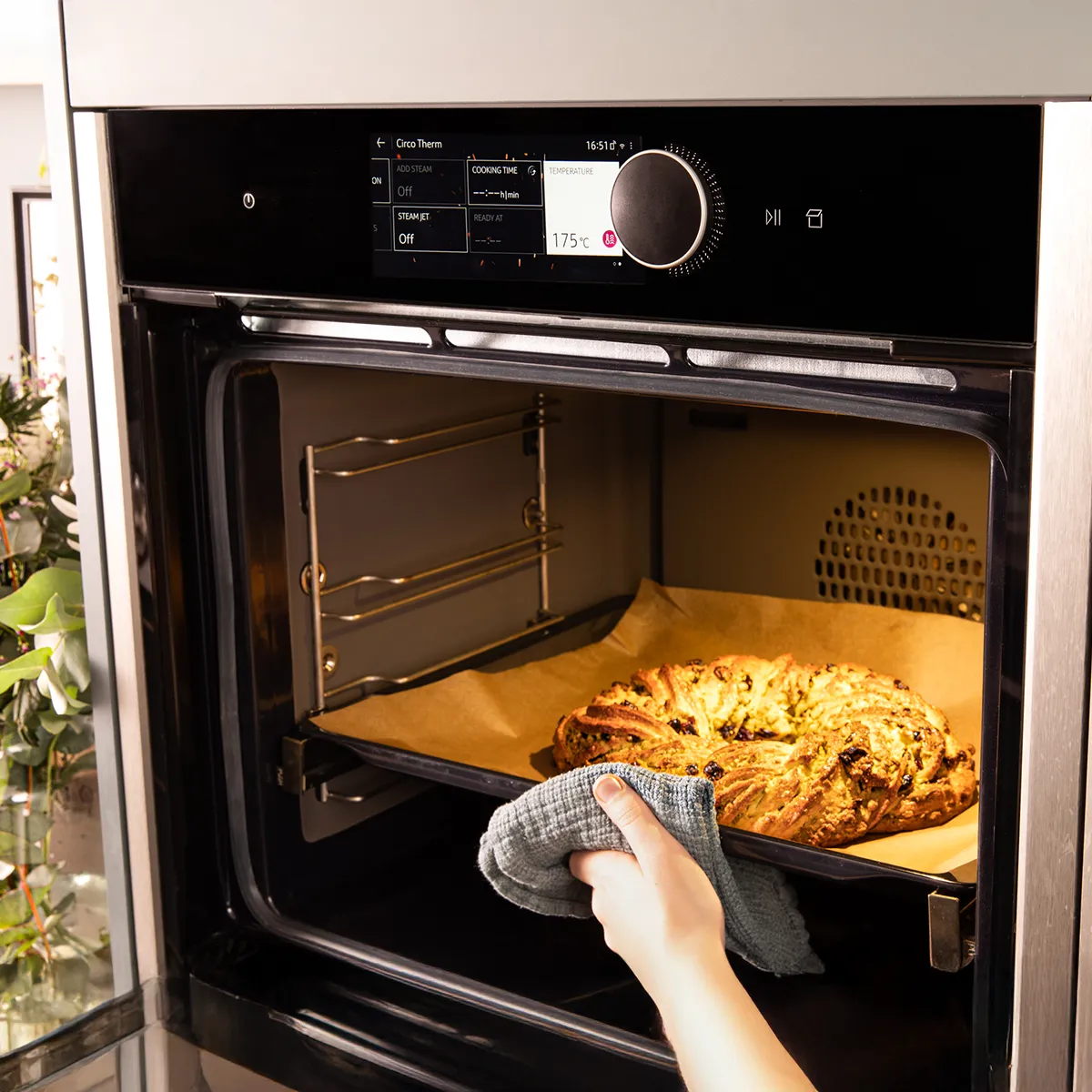 A hand in a grey mitt pulls a golden braided bread with dried fruit from a modern oven displaying 175°C.