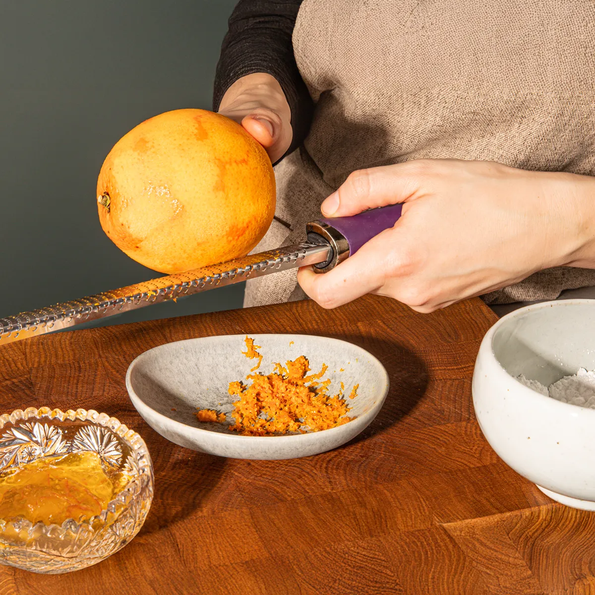 A person grating an orange over a small bowl, with zest accumulating in the bowl and a dish of honey nearby on a wooden table.