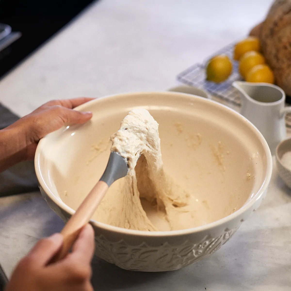 Une personne remue une pâte épaisse dans un bol en céramique, avec des citrons et du pain cuit visibles en arrière-plan.