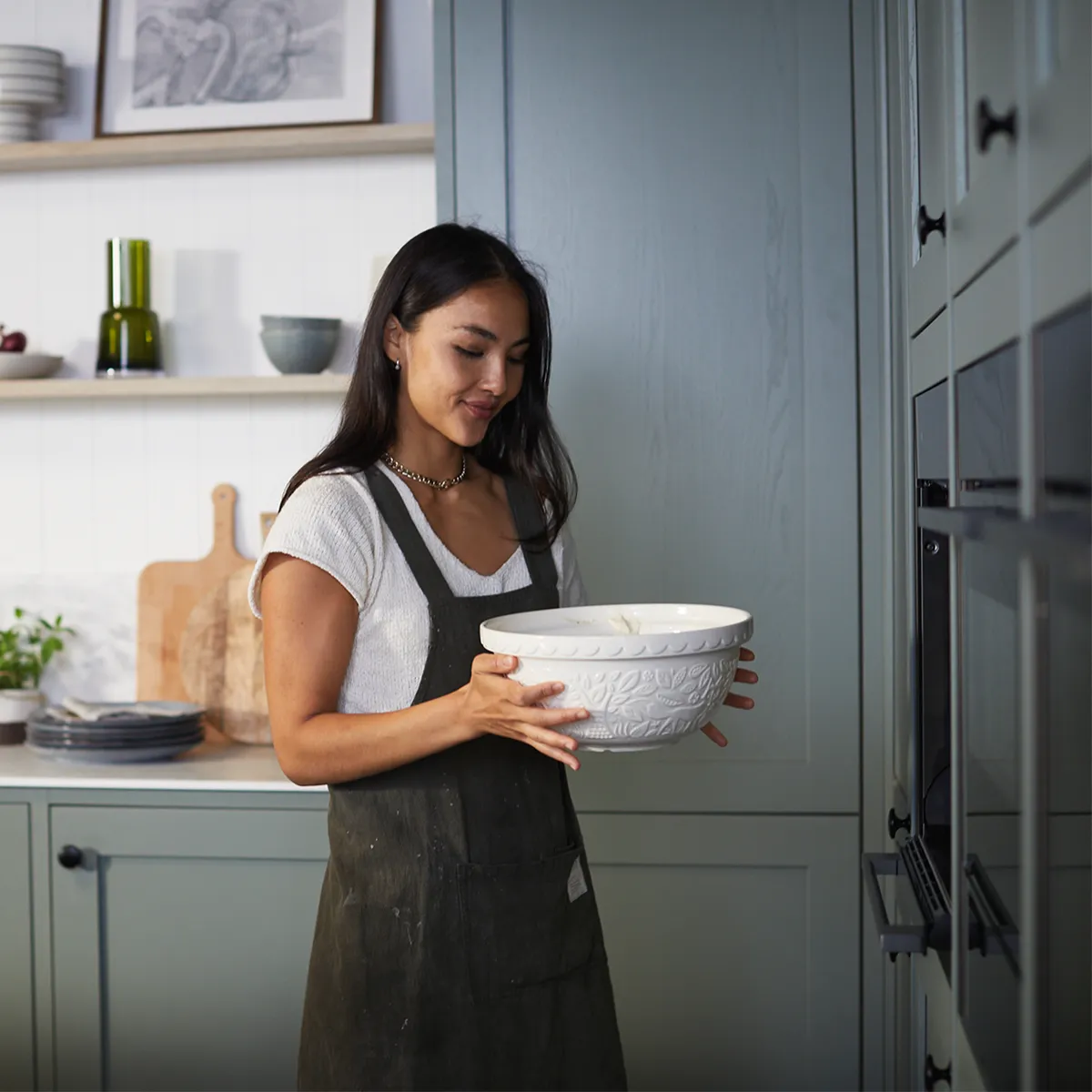 Dans une cuisine moderne, une personne en tablier tient un grand bol décoratif. Des étagères avec des ustensiles de cuisine et des plantes sont visibles en arrière-plan.