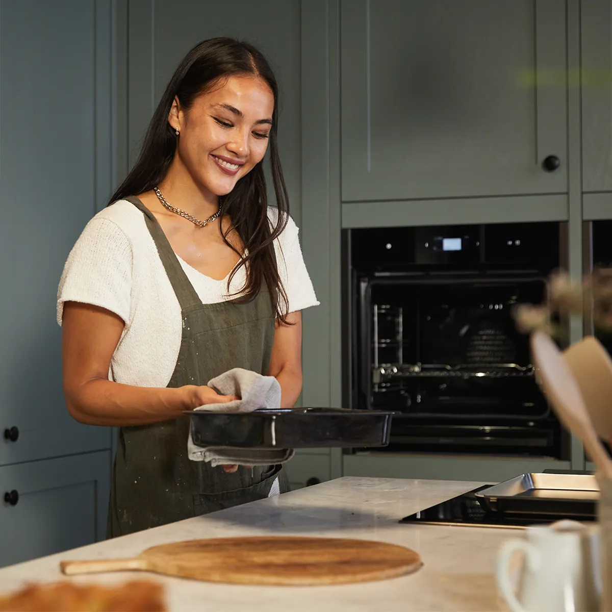 Femme souriante en tablier vert tenant un plat de cuisson dans une cuisine élégante avec des armoires vertes et un four encastré.