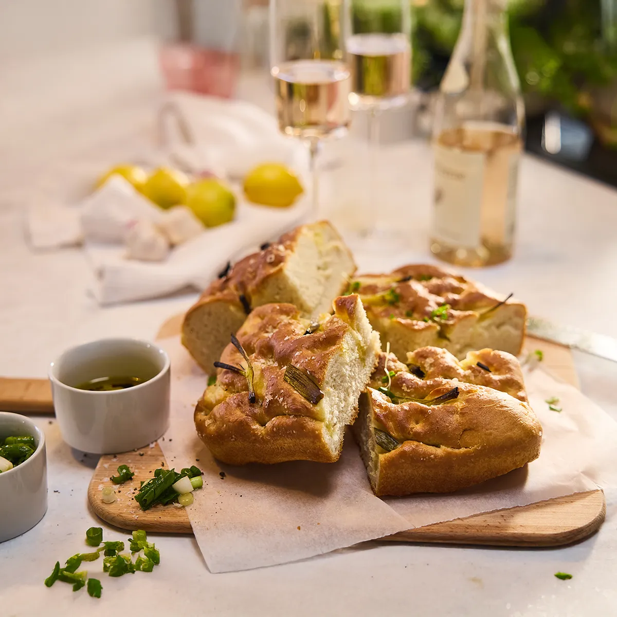 Pain focaccia tranché avec de l'huile d'olive et des oignons verts sur une planche, avec du vin, des citrons et de l'ail en arrière-plan.