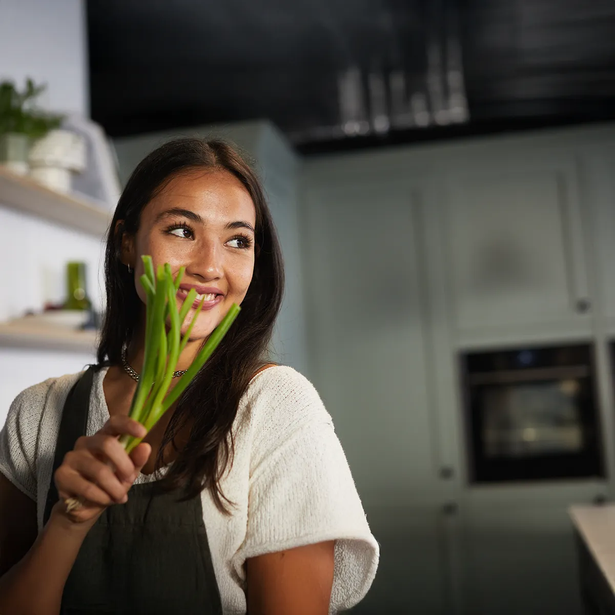 Femme souriante dans la cuisine, tenant des poireaux verts et regardant sur le côté.