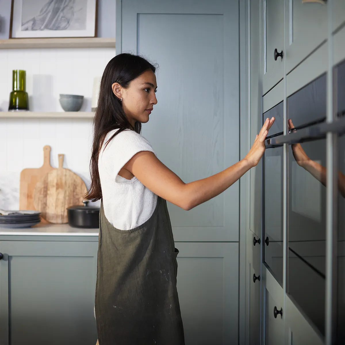 Femme en tablier vert touchant un four encastré dans une cuisine élégante avec des armoires vertes.