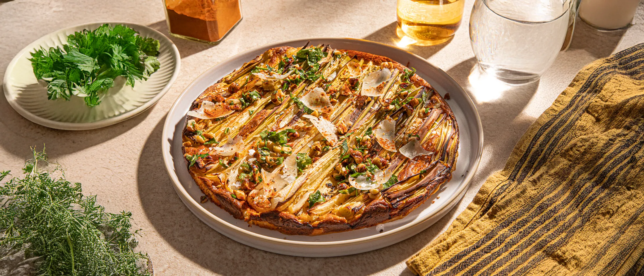 A beautifully arranged savory tart topped with green onions, herbs, and cheese, surrounded by fresh cilantro and a glass of water.