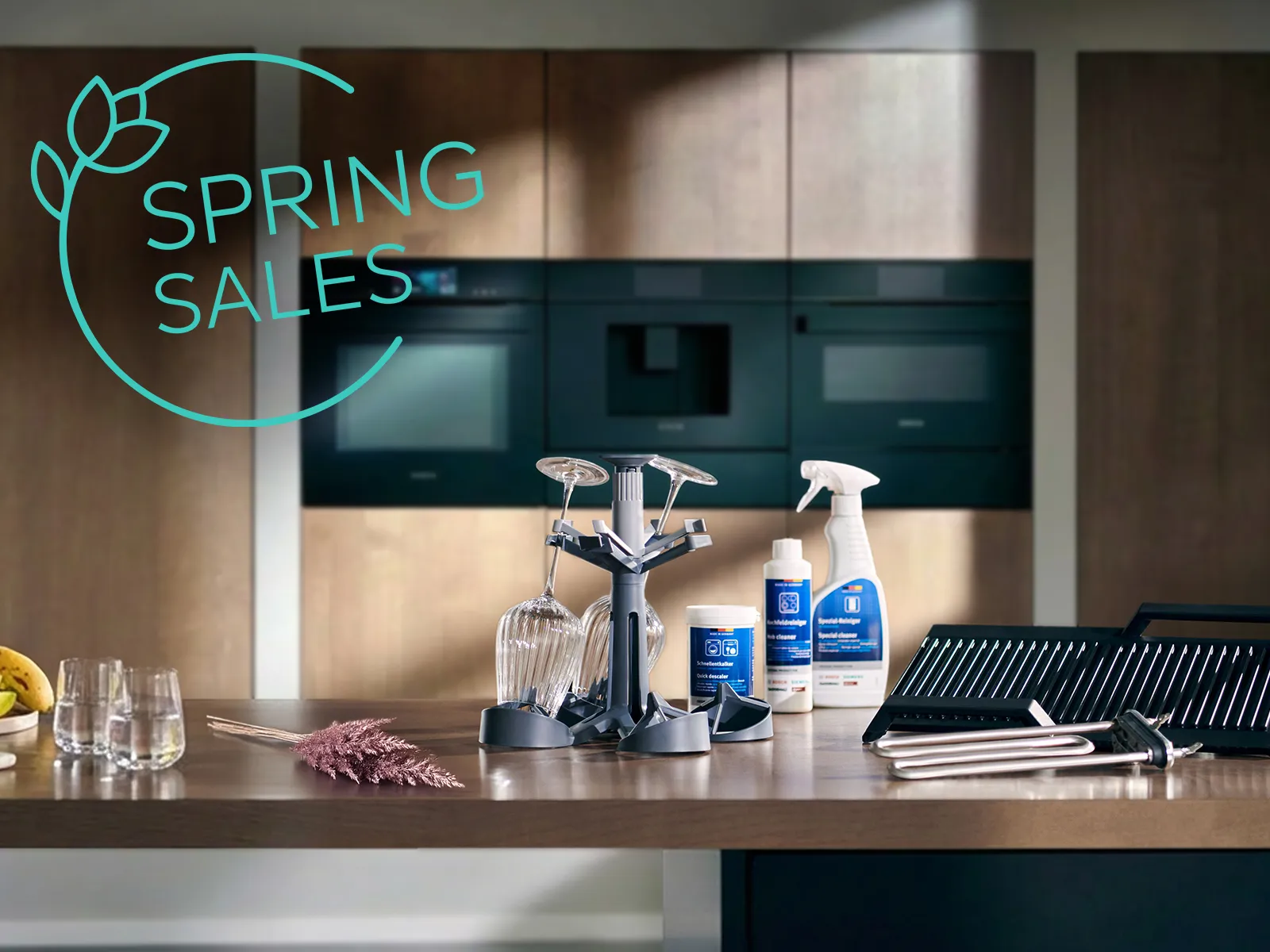 Modern kitchen countertop with cleaning products, dish rack, fruit bowl, glasses, and a woman walking near a window; "Spring Sales" text overlay.