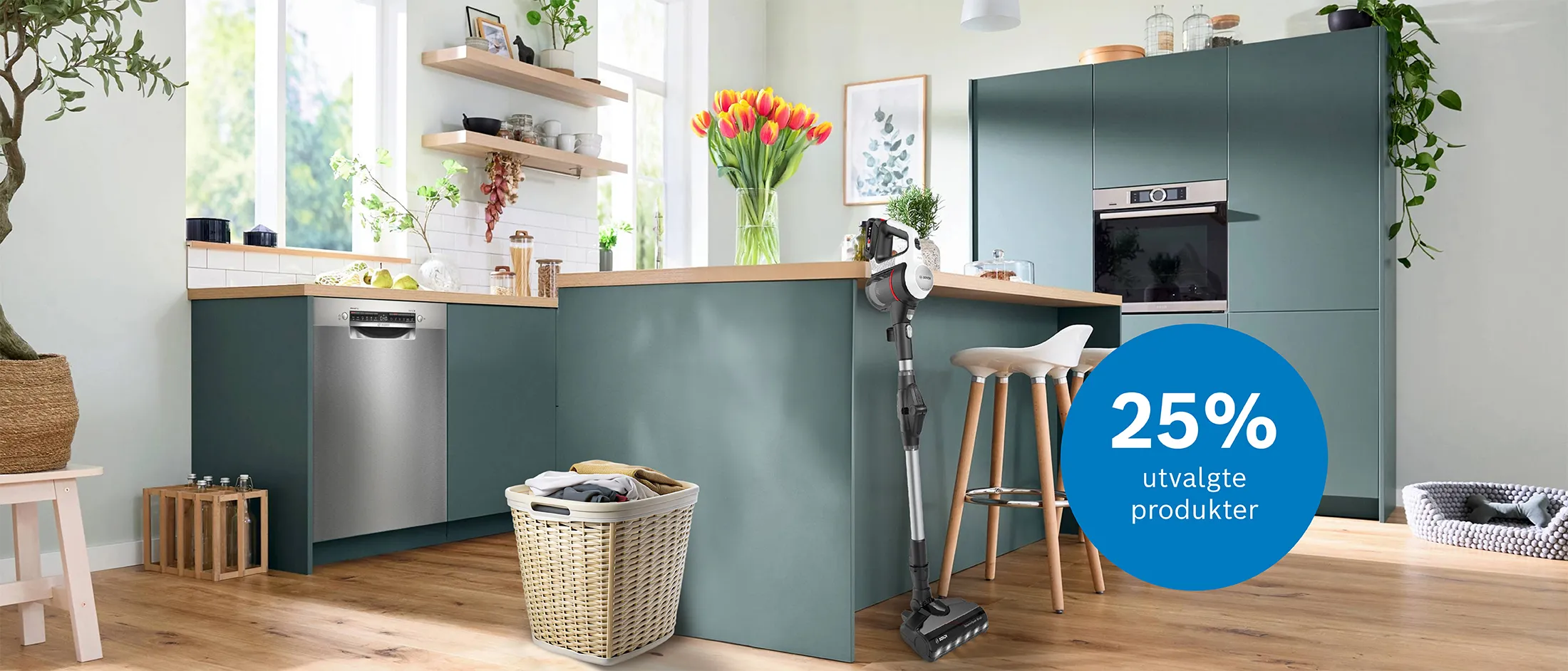 Et blågrønt kjøkken med lyse tredetaljer, en bukett med tulipaner på kjøkkenøya, barstoler i lyst tre, en skittentøyskurv står foran kjøkkenøya, og en Unlimited-støvsuger lener seg mot kjøkkenøya. I bakgrunnen sees ovn og oppvaskmaskin som er pent integrert i kjøkkenet.