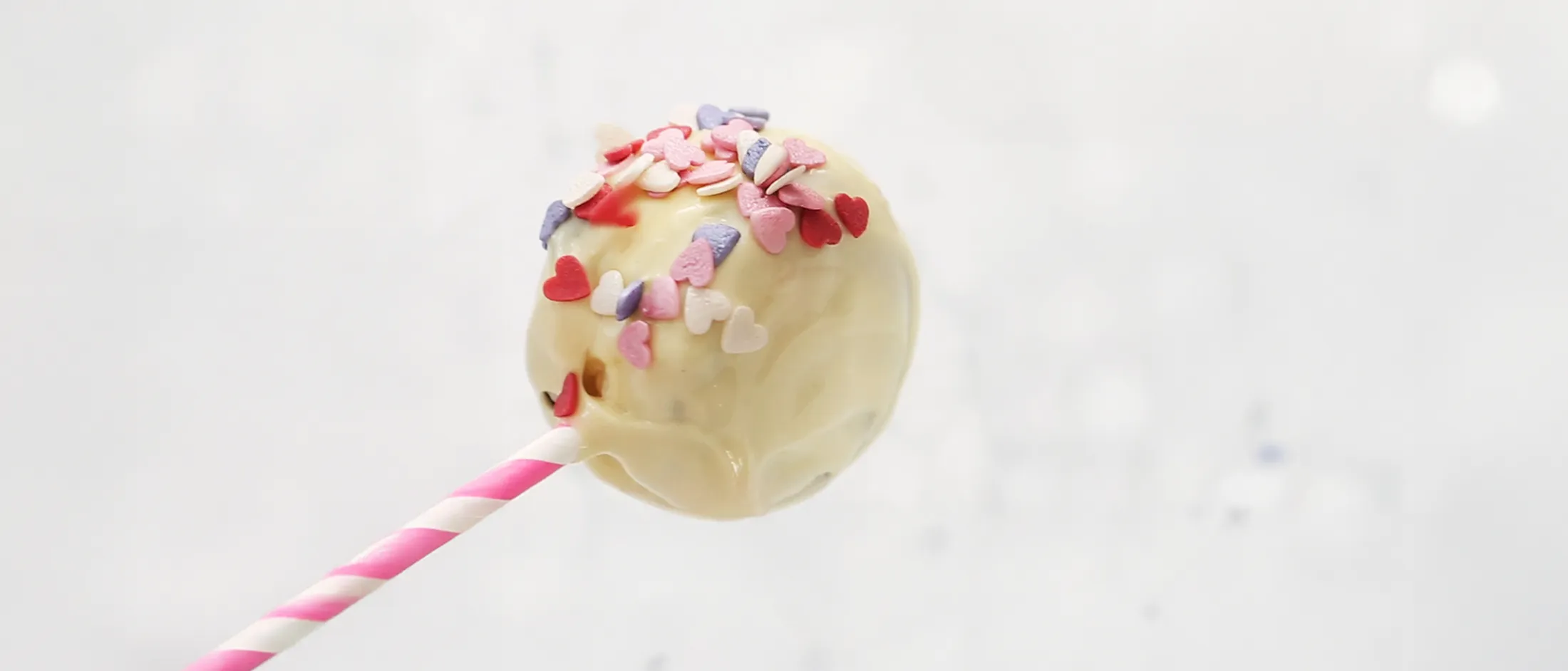 A hand decorating a white chocolate-covered cake pop with colorful heart-shaped sprinkles.