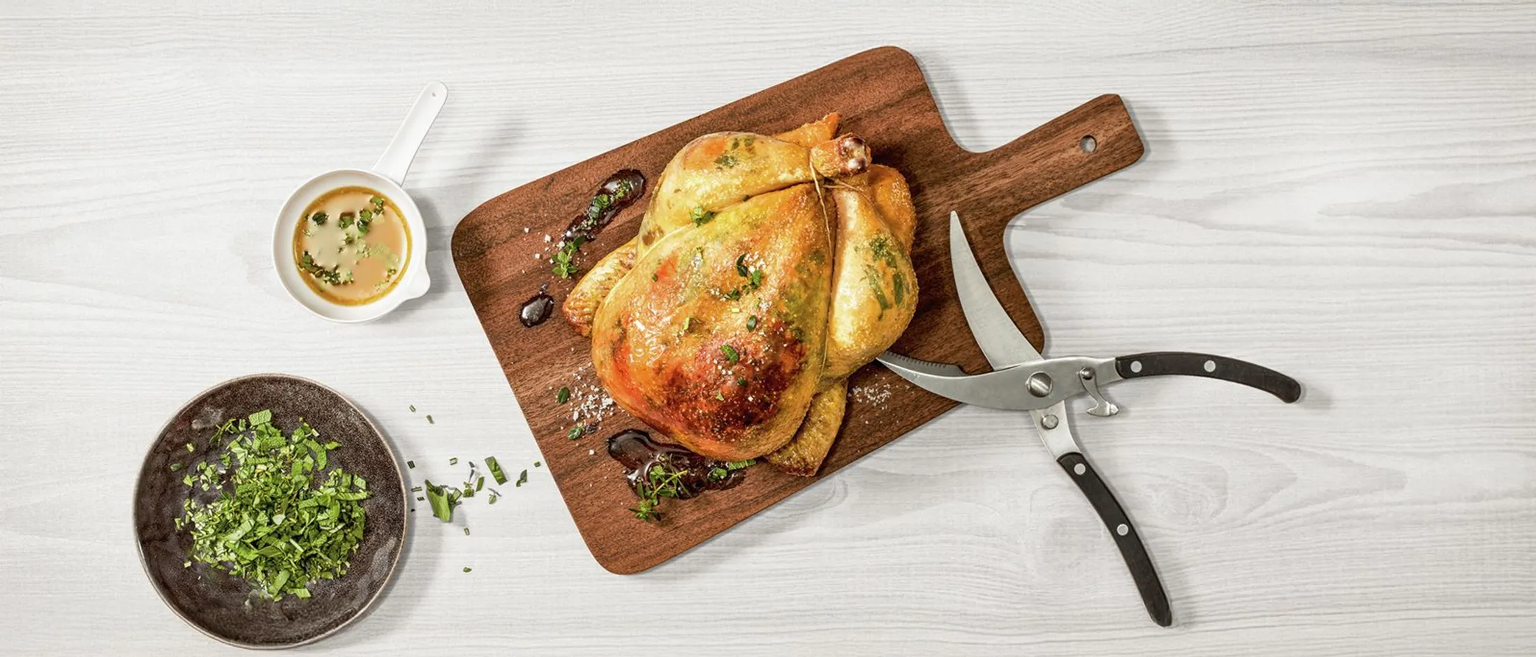 Whole roasted chicken on a wooden cutting board with herb sauce, chopped fresh herbs on a plate, and kitchen shears on a light wood table.