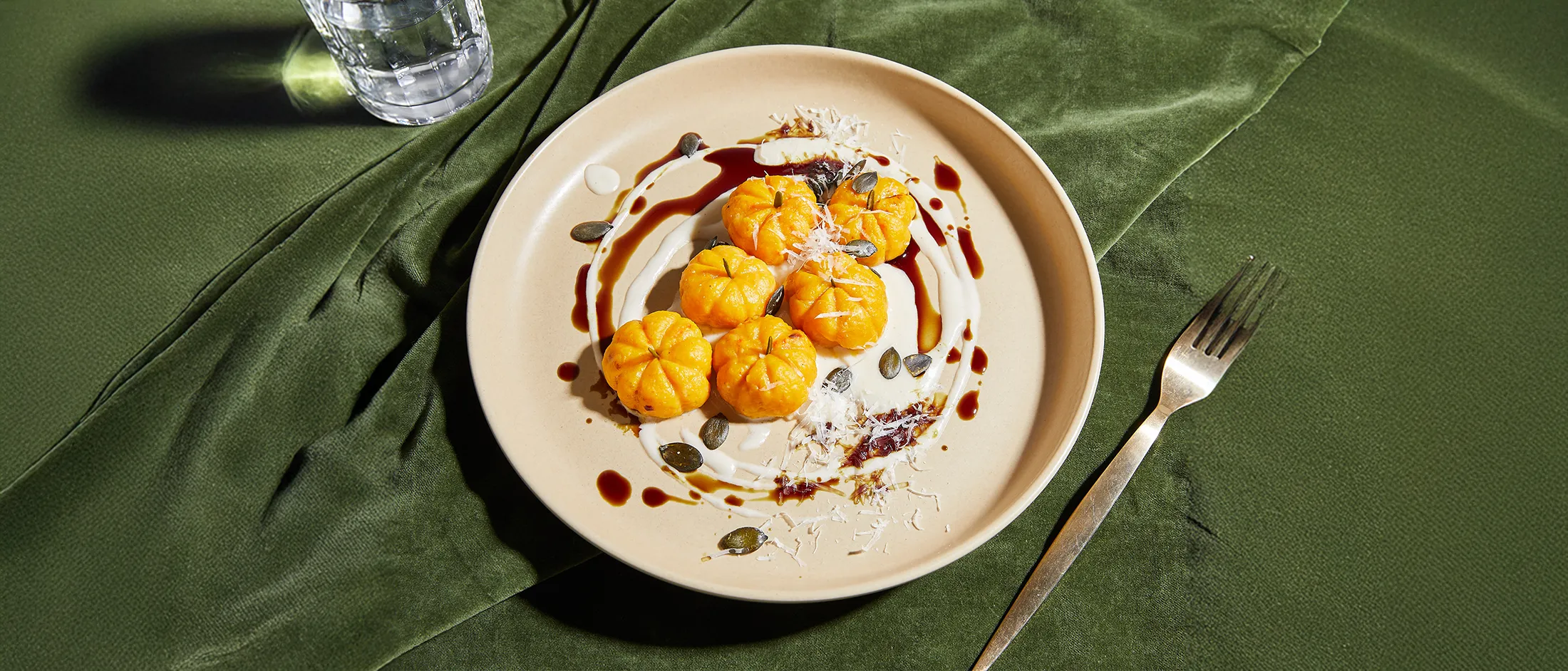 Plate of six pumpkin-shaped gnocchi with cream sauce, balsamic glaze, pumpkin seeds, and grated cheese on a beige plate with a green cloth background.