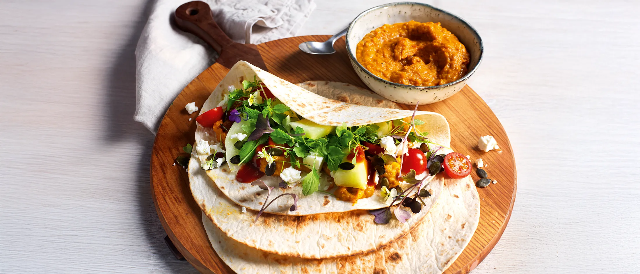 Soft tortilla wrap filled with fresh greens, cherry tomatoes, cheese, and a bowl of thick orange dip on a wooden serving board