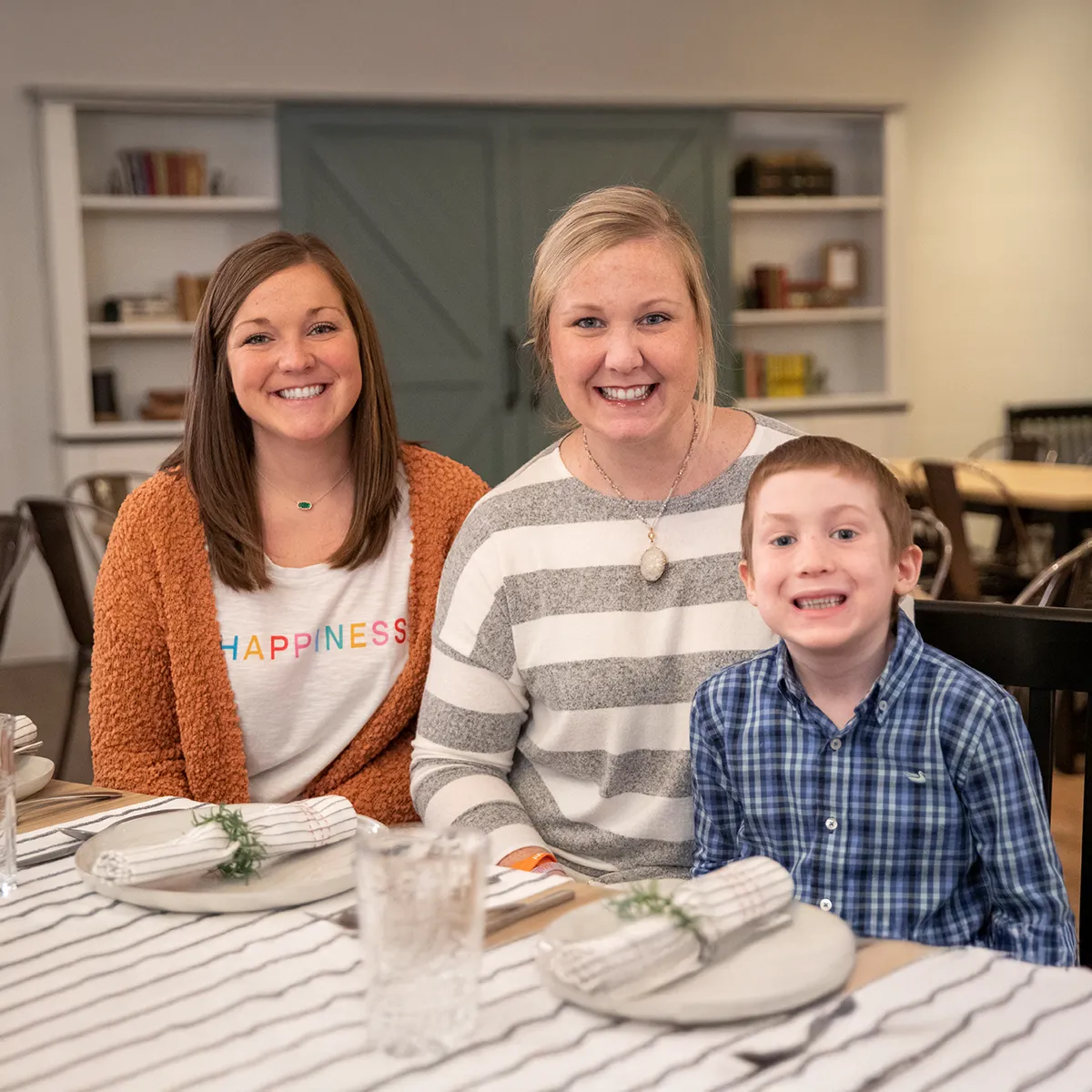 Three people sitting at a dining table set with plates, napkins, and glasses in a cozy room with shelves and a green door in the background.