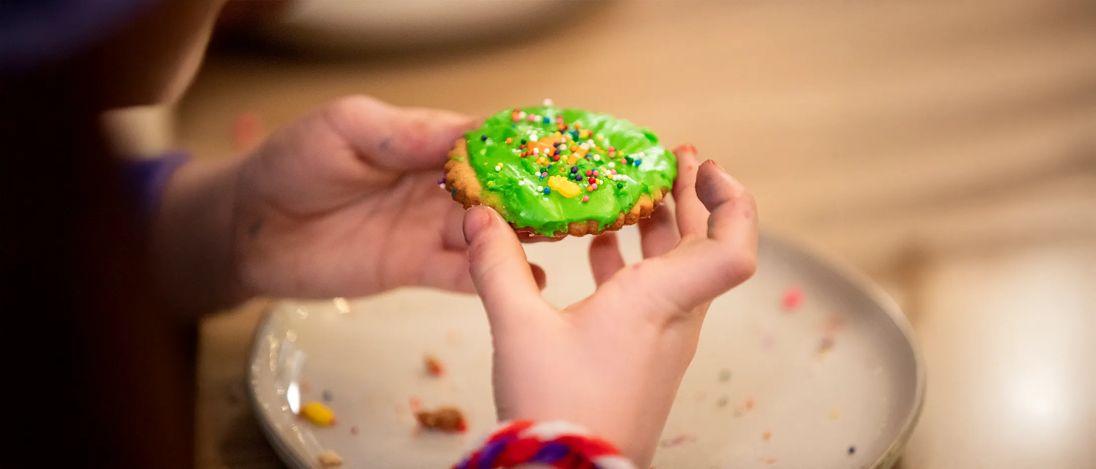 Child's hands holding a round cookie with green frosting and colorful sprinkles over a white plate.