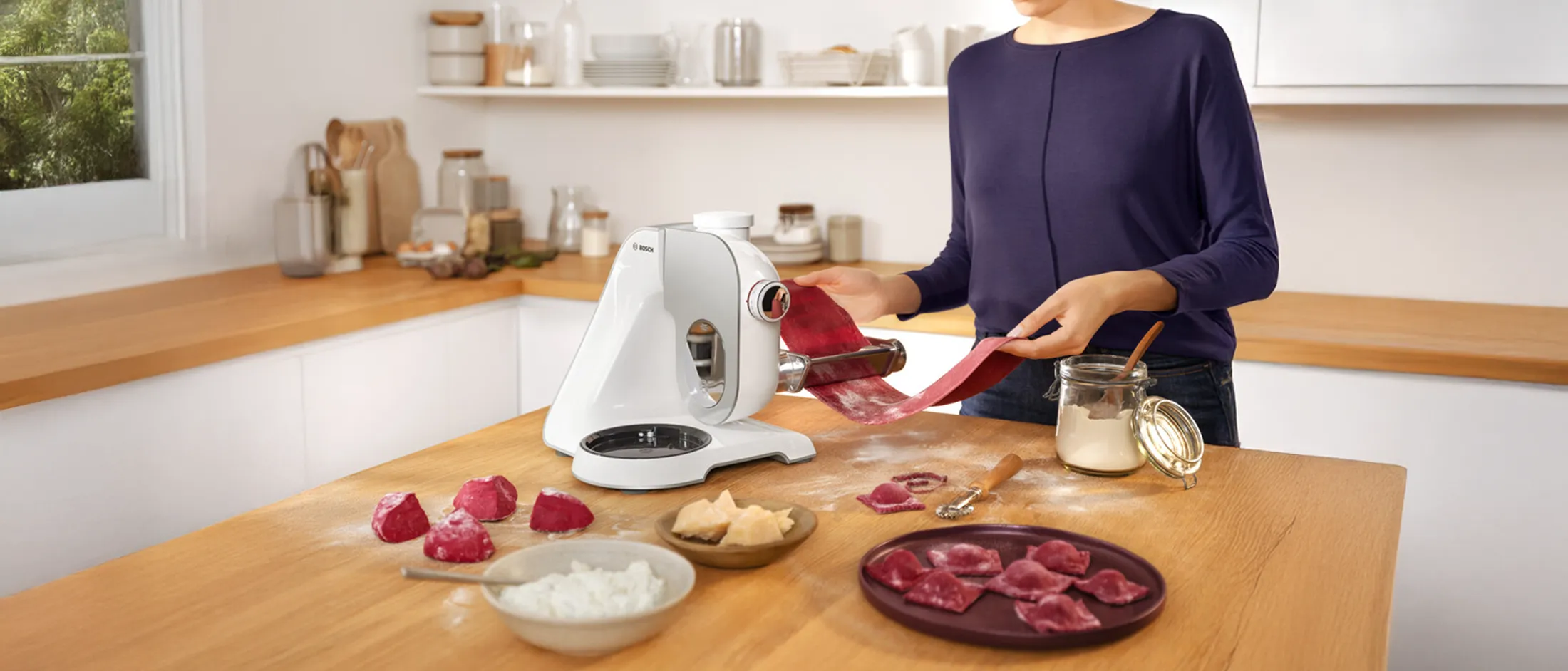 Person using a pasta machine to roll out red dough on a wooden kitchen counter with ingredients and prepared ravioli nearby.