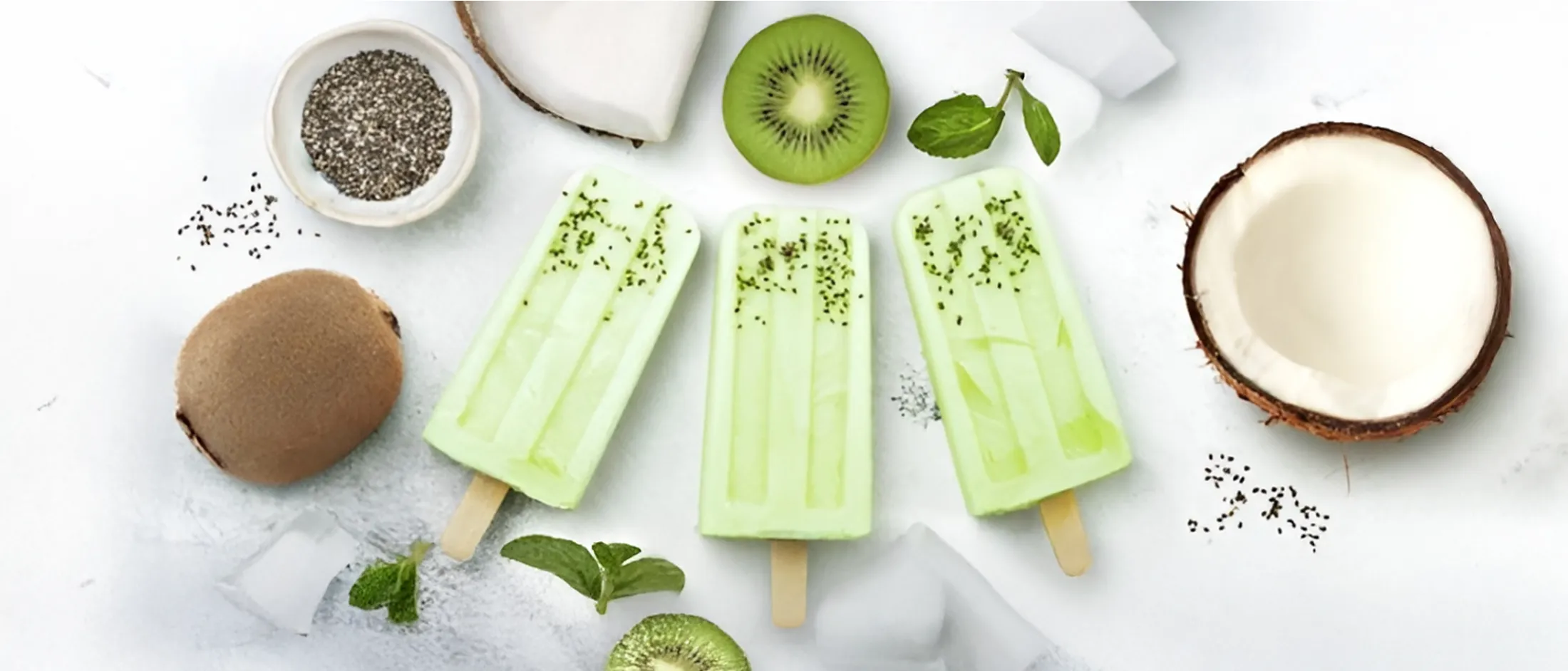 Three green kiwi popsicles with chia seeds on ice, surrounded by kiwi slices, coconut halves and mint leaves.