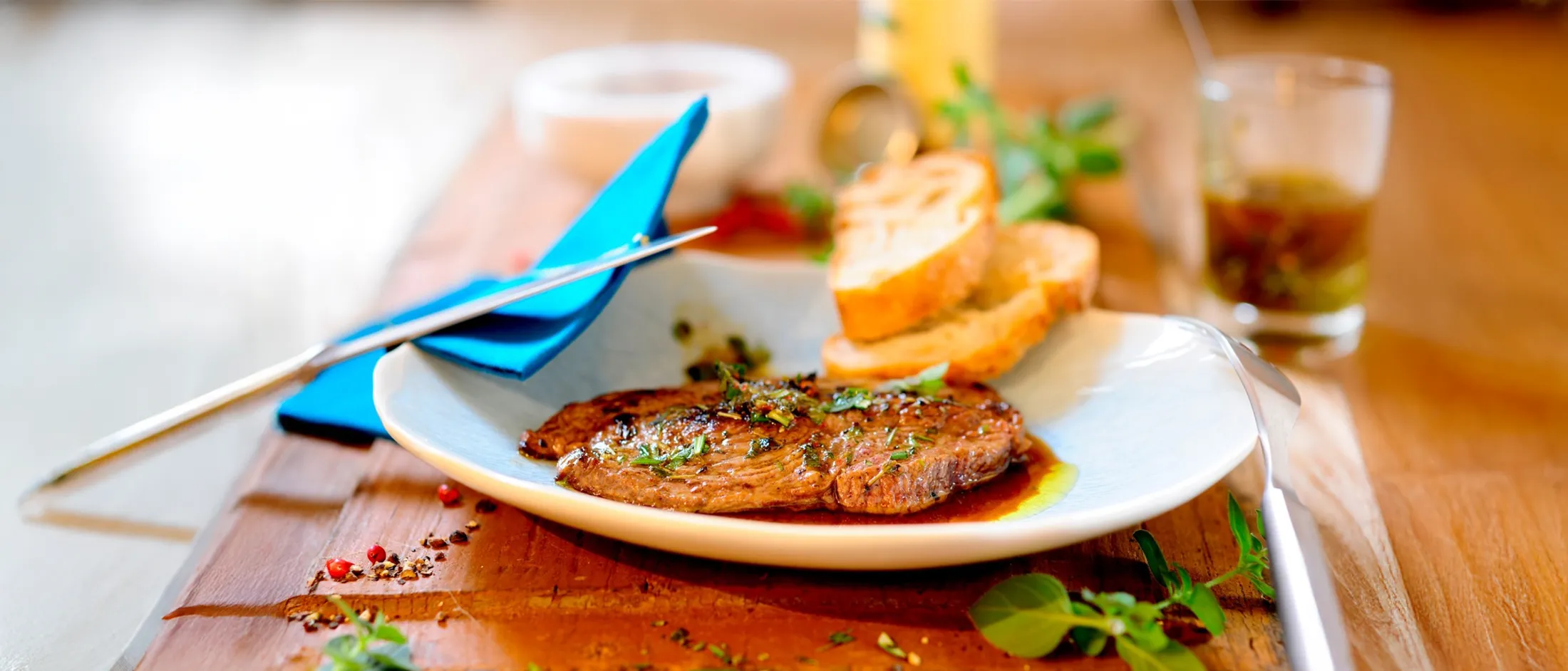 Grilled steak topped with chopped herbs and sauce on a white plate, served with two slices of toasted baguette, a blue napkin, and scattered herbs on a wooden table.