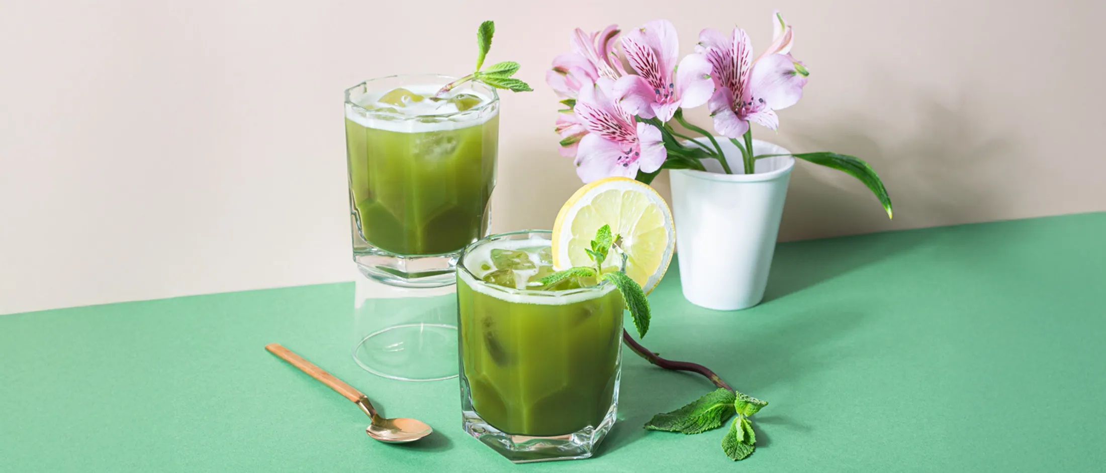Two iced green drinks garnished with mint and lemon, a small bouquet of pink flowers in a white vase, and a gold spoon on a green surface.